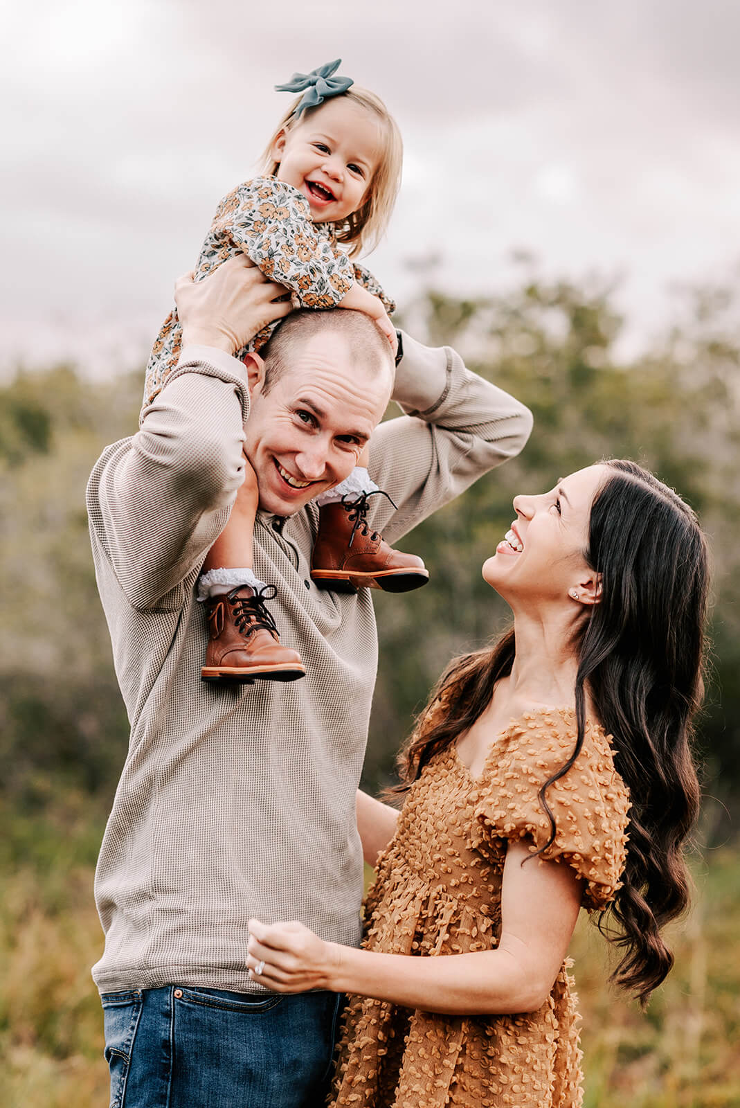 A dad plays with his daughter on his shoulders with mom laughing with them in a park after visiting rollys baby boutique
