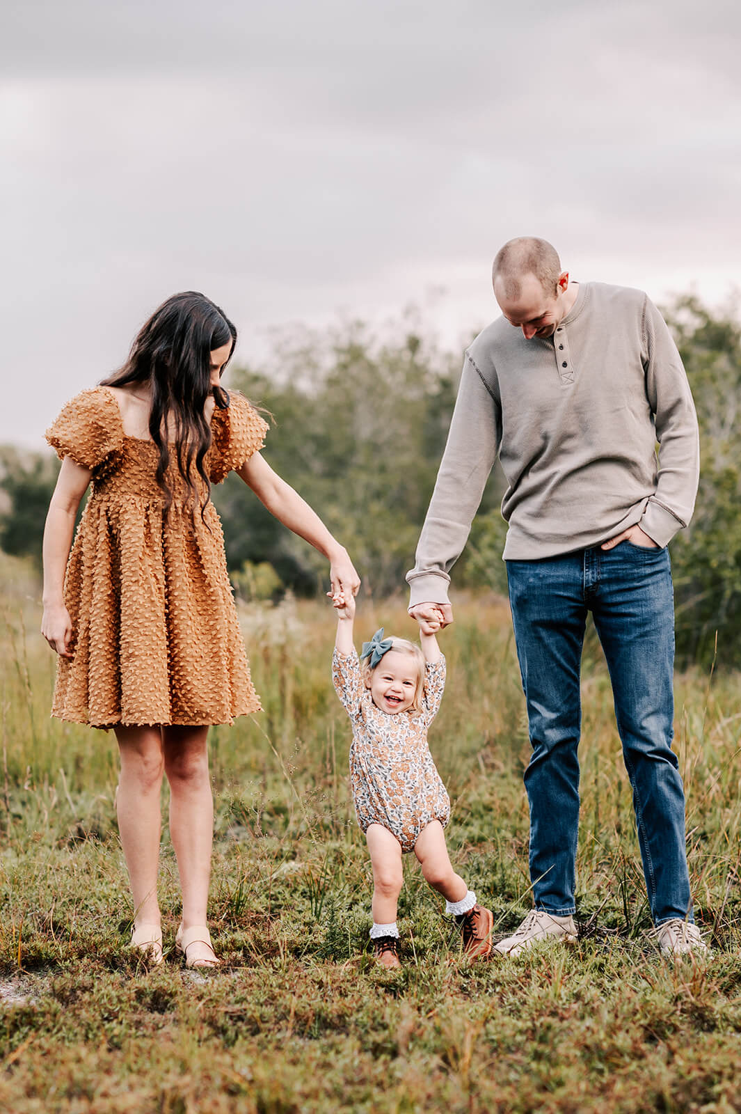 A mom and dad hold hands with their toddler daughter while walking through a grassy field after visiting toads and tulips furniture