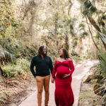 A mom to be in. red maternity gown walk down a forest sidewalk smiling with her husband