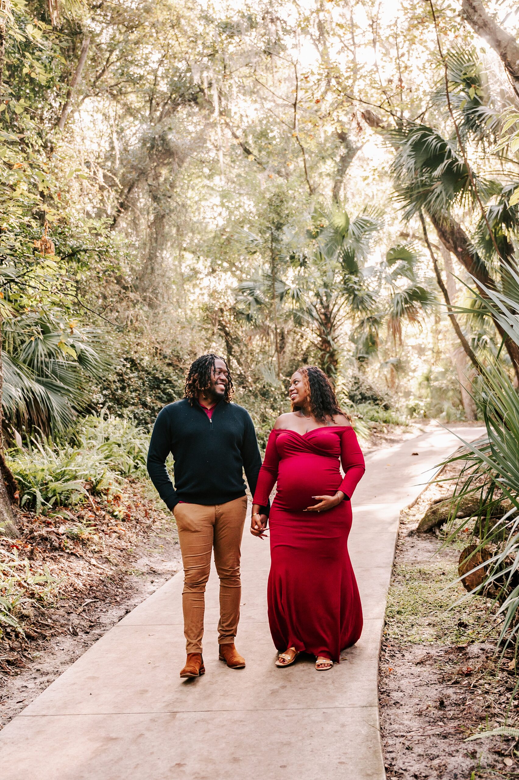 A mom to be in. red maternity gown walk down a forest sidewalk smiling with her husband