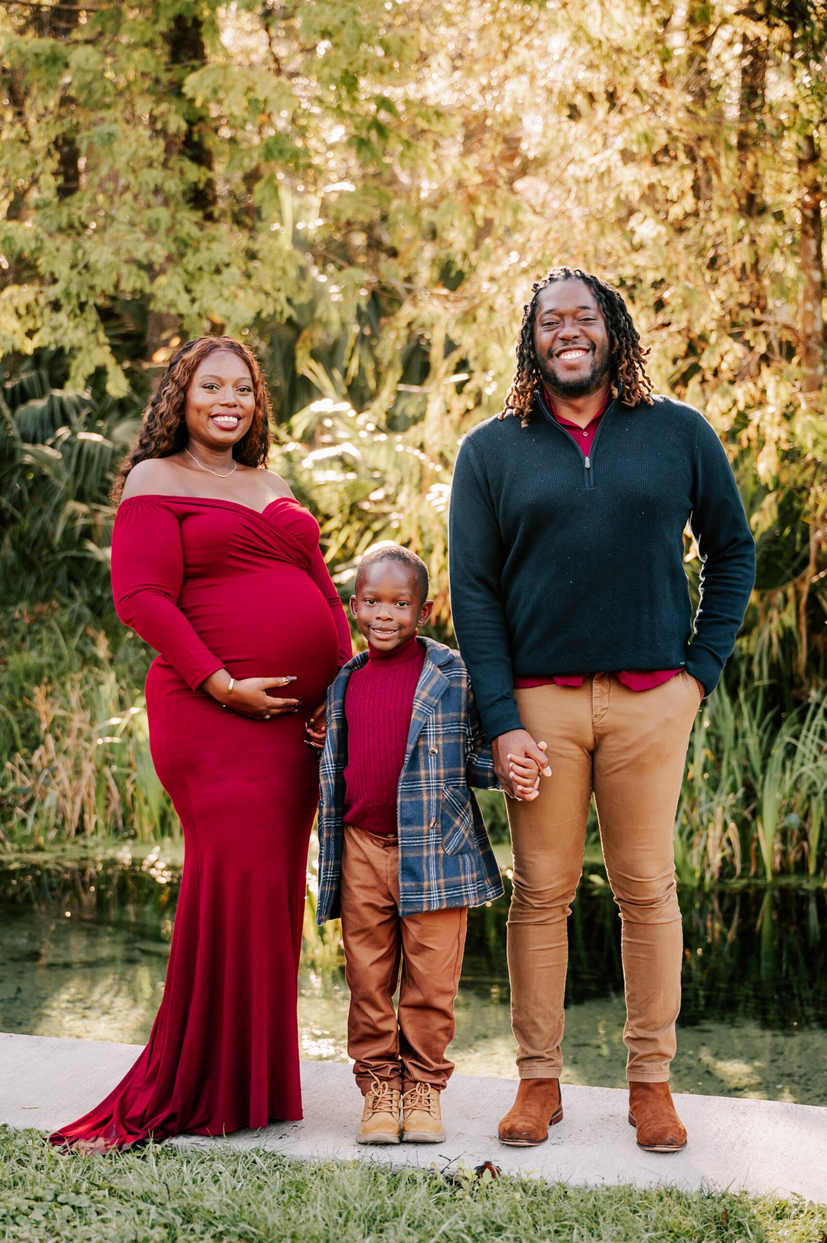 A pregnant mother stands on a riverside sidewalk holding hands with her young son and husband in a red maternity gown after meeting triangle doulas of color