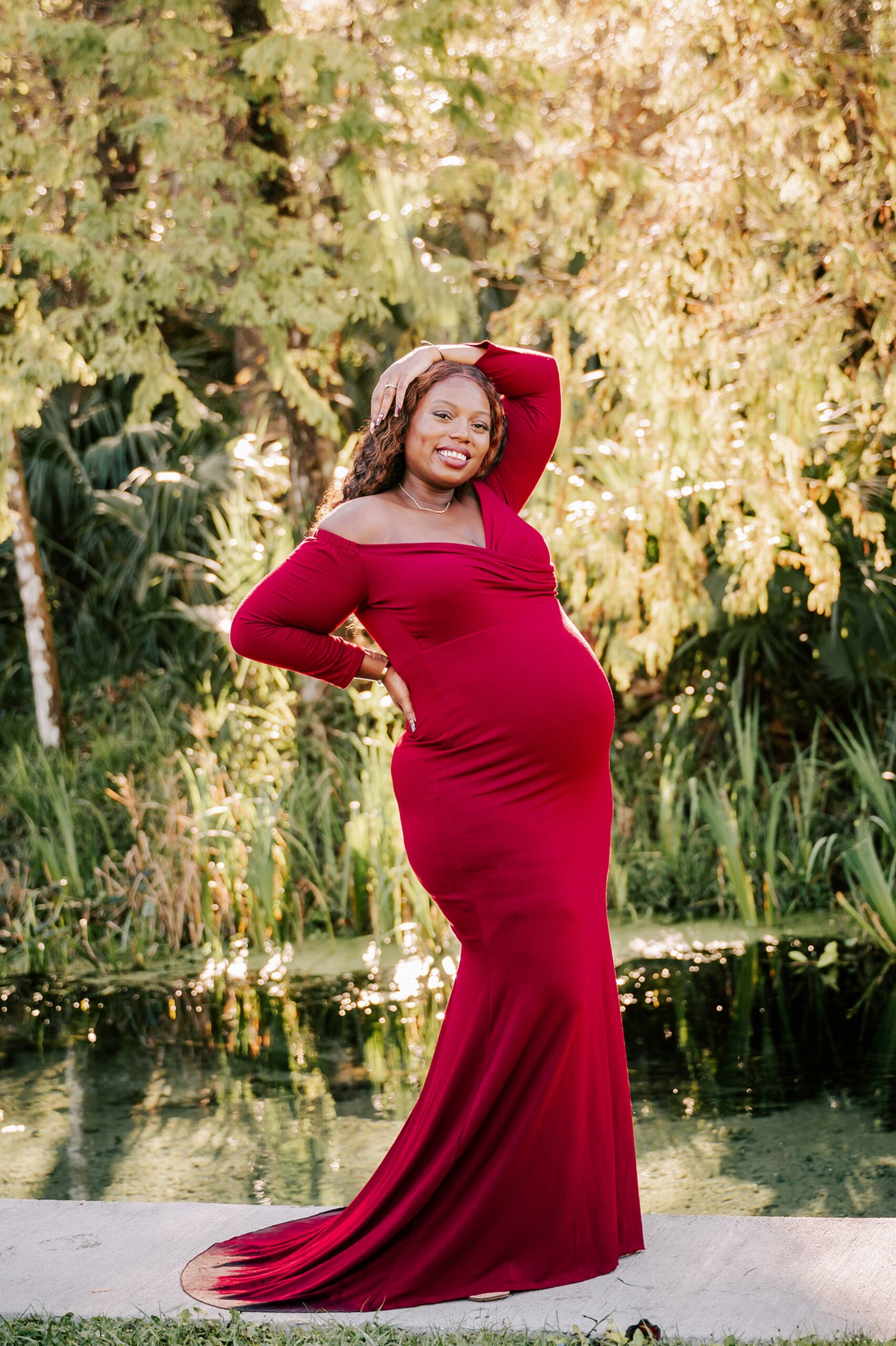 A mother to be stands on the edge of a river in a long red maternity gown after meeting triangle doulas of color