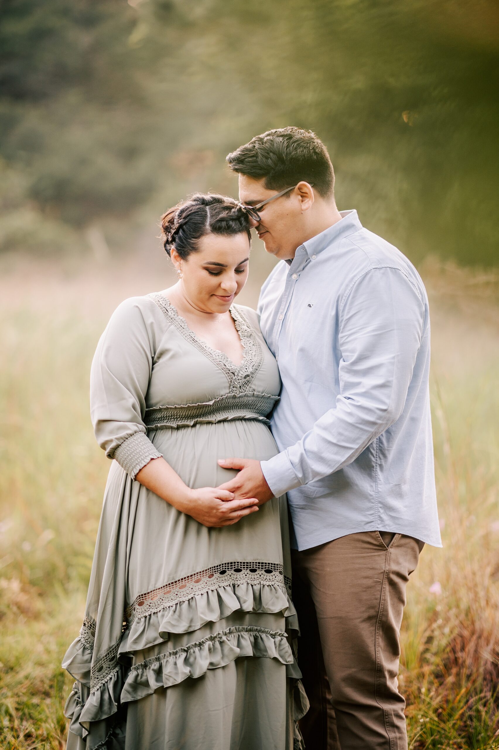 A mother to be in a green maternity dress stands close with her husband in a grassy green field at sunset holding the bump