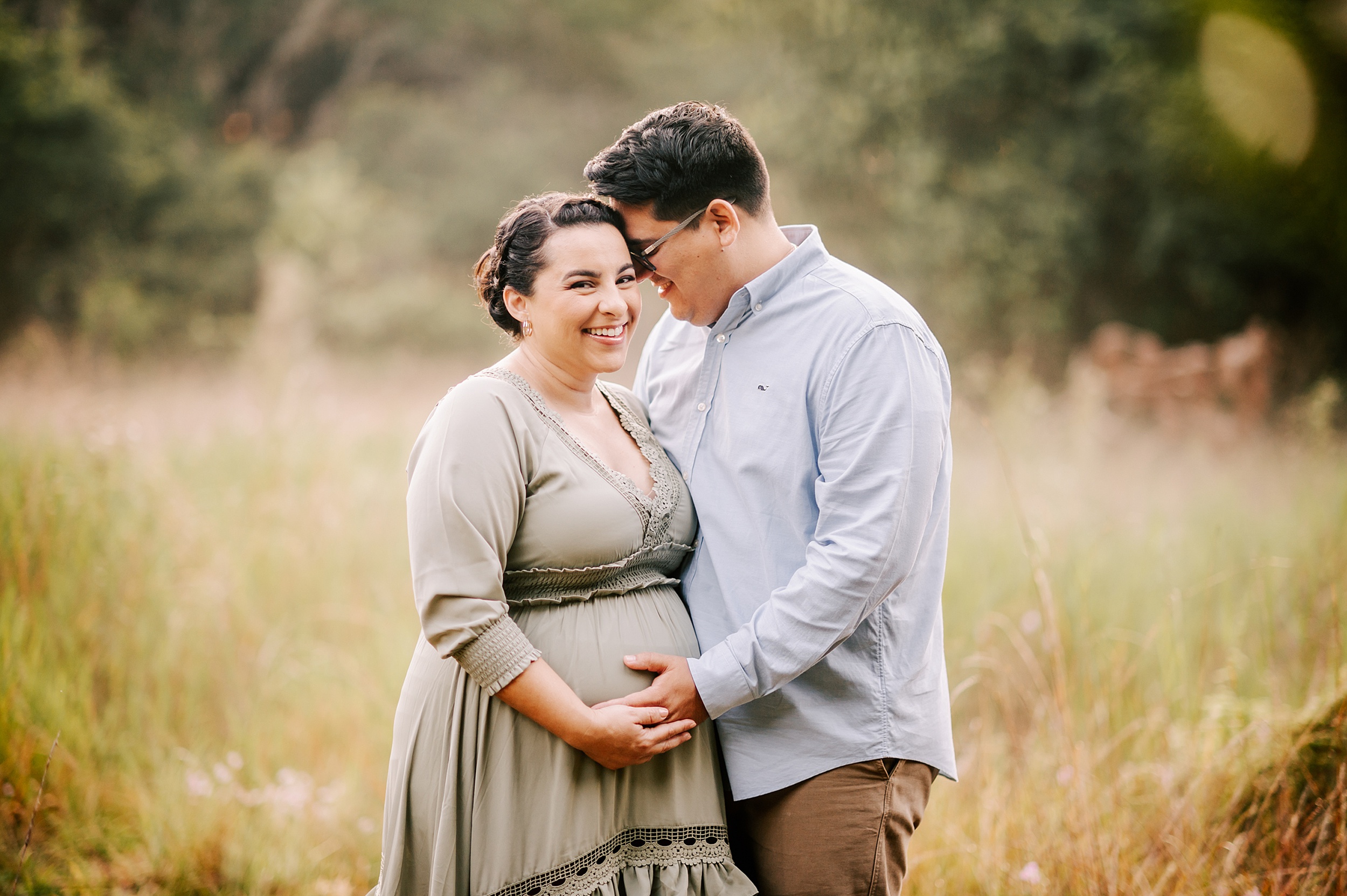 A mother to be laughs with her husband as they stand in a field of tall grass holding the bump after visiting triangle mothercare winston salem