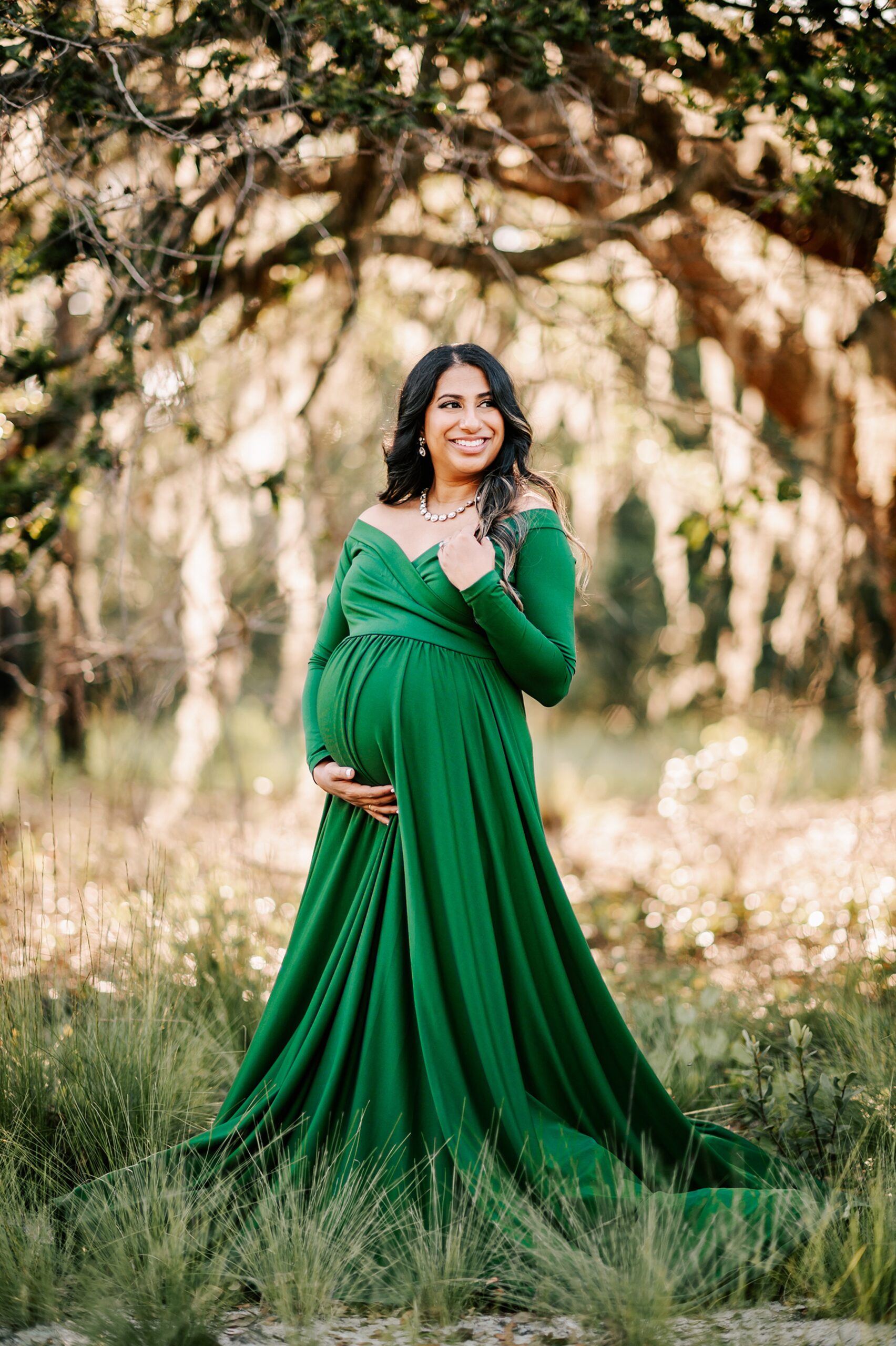 A mother to be wearing a long green maternity gown stands in a forest at sunset