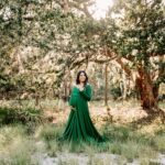 A mother to be in a green maternity gown smiles over her shoulder while standing in a forest at sunset after visiting unc birth center
