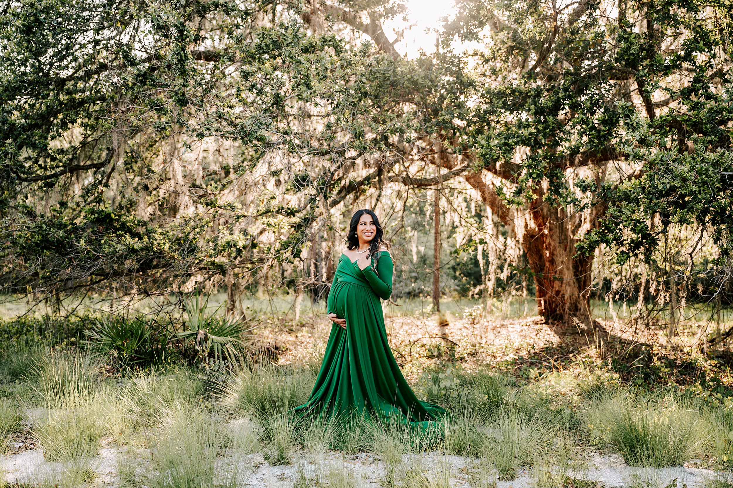 A mother to be in a green maternity gown smiles over her shoulder while standing in a forest at sunset after visiting unc birth center