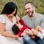 A mom and dad play and laugh with their toddler daughter in dad's lap while sitting on a blanket in a park after using wake forest obgyn clemmons