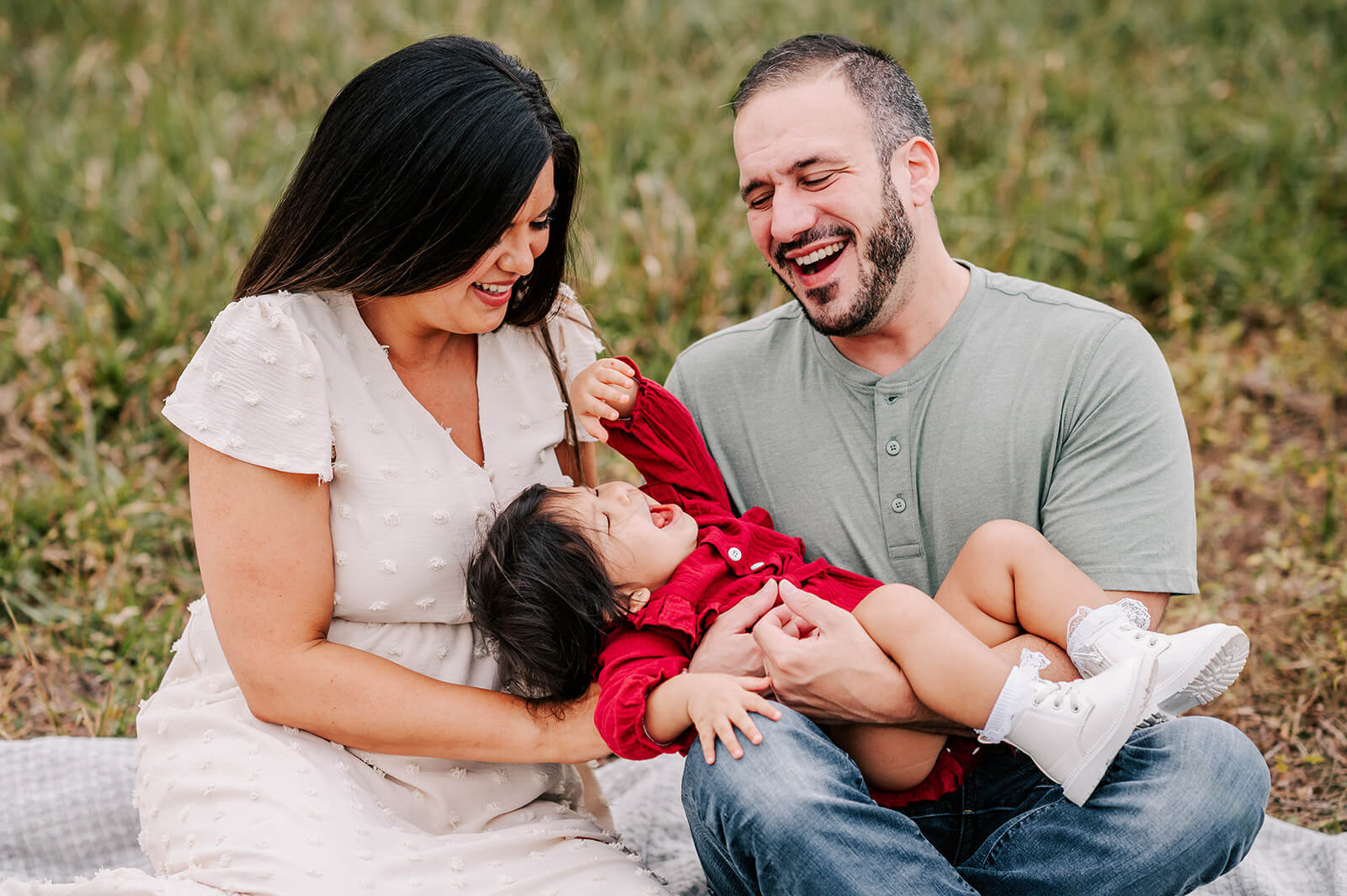 A mom and dad play and laugh with their toddler daughter in dad's lap while sitting on a blanket in a park after using wake forest obgyn clemmons