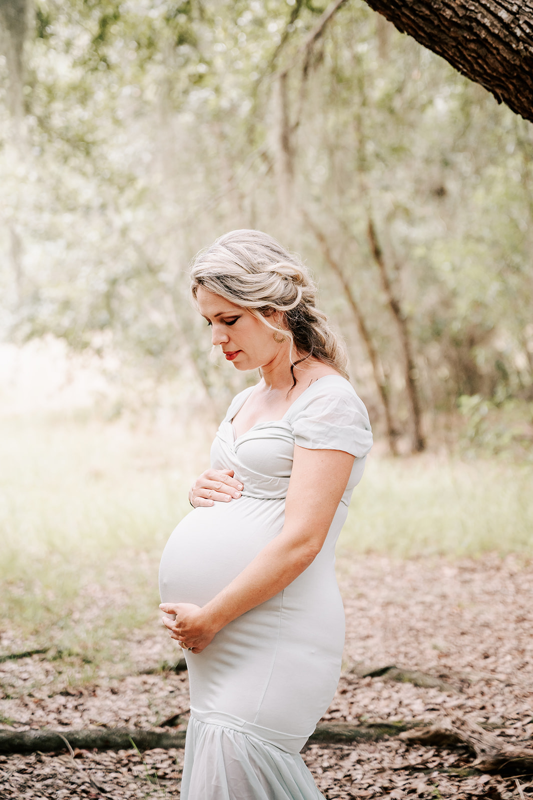 A mother to be holds her bump while standing in a forest in a baby blue maternity gown