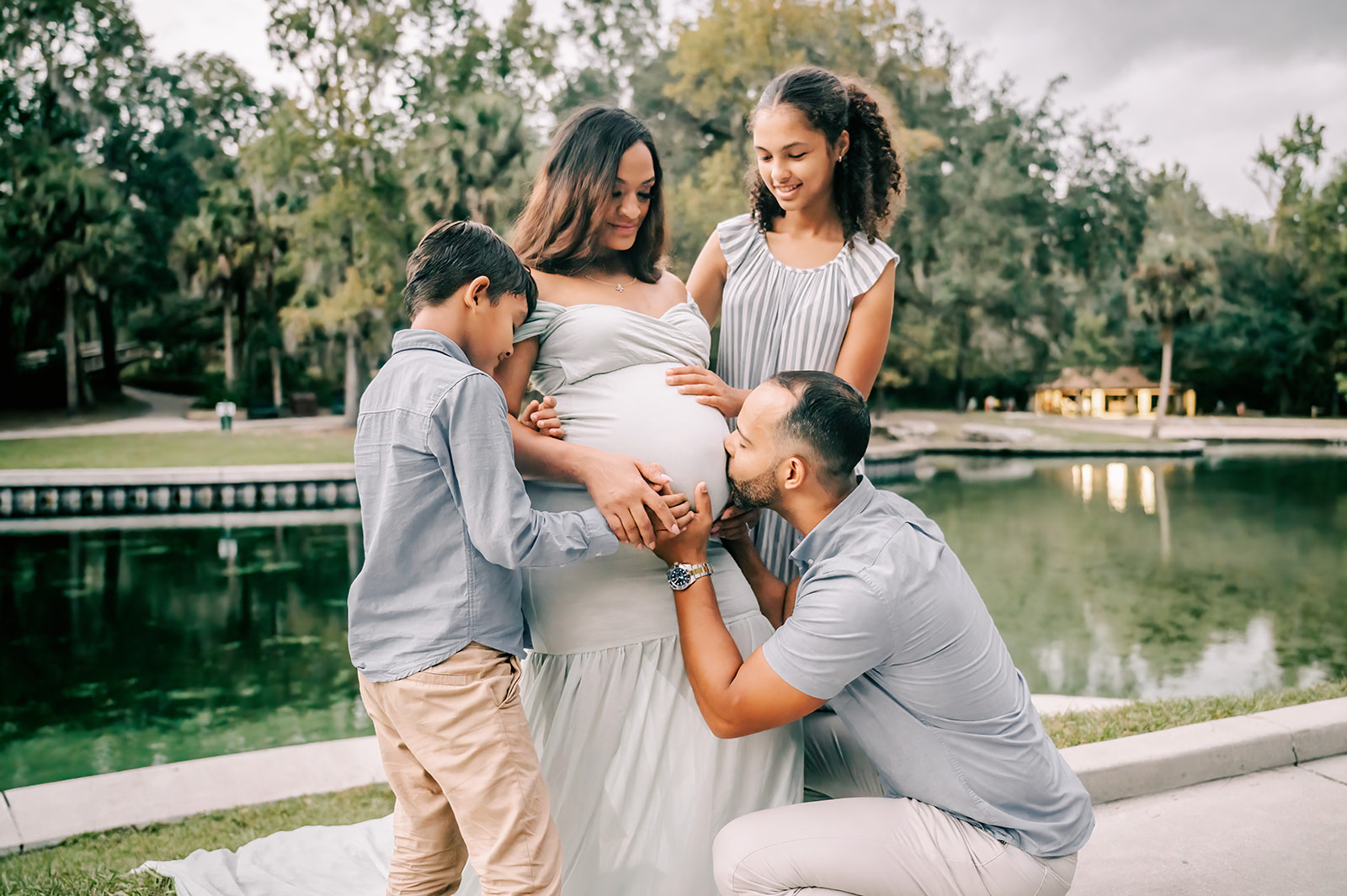 A pregnant mother in a blue maternity gown stands on a river sidewalk while dad kisses the belly and her two young children hold the bump after visiting Mindful Family Wellness