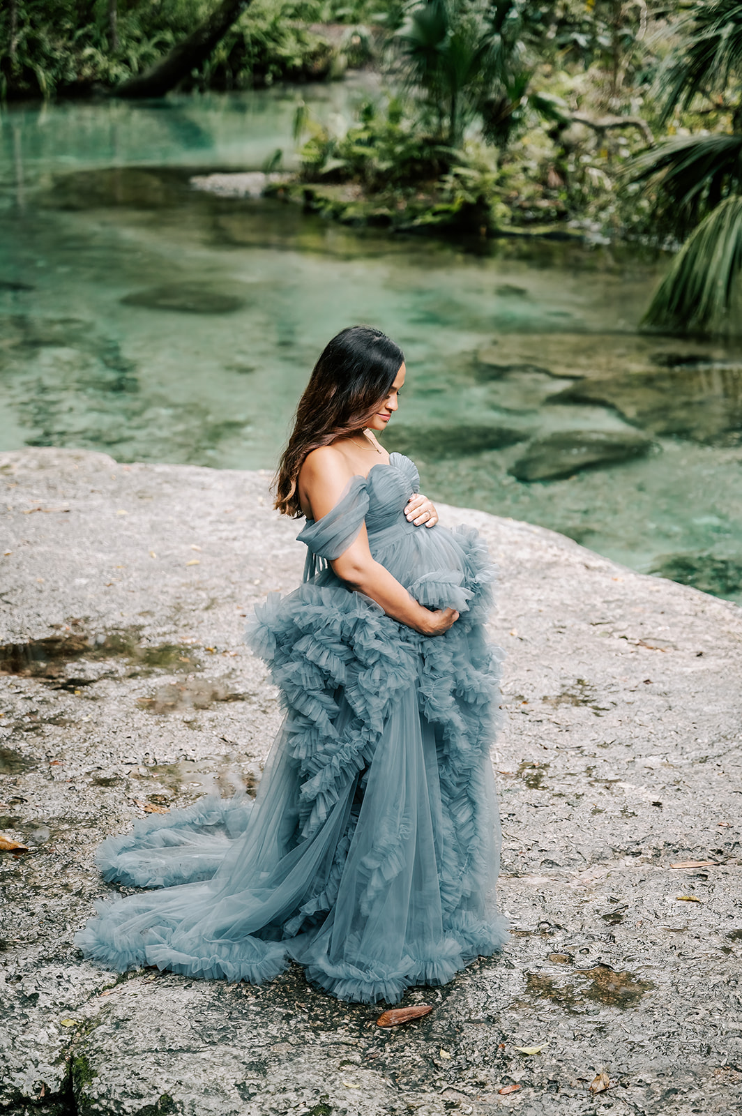 A mother to be smiles down to her bump while standing in the springs in a blue maternity gown
