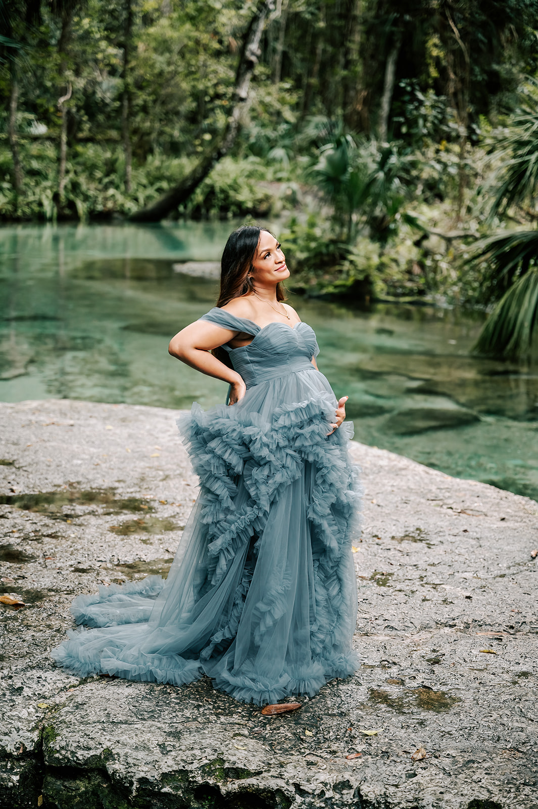 A mom to be smiles while holding her bump in a long blue maternity gown and standing on a large rock on a clear river after visiting Moonshadow Medical Massage