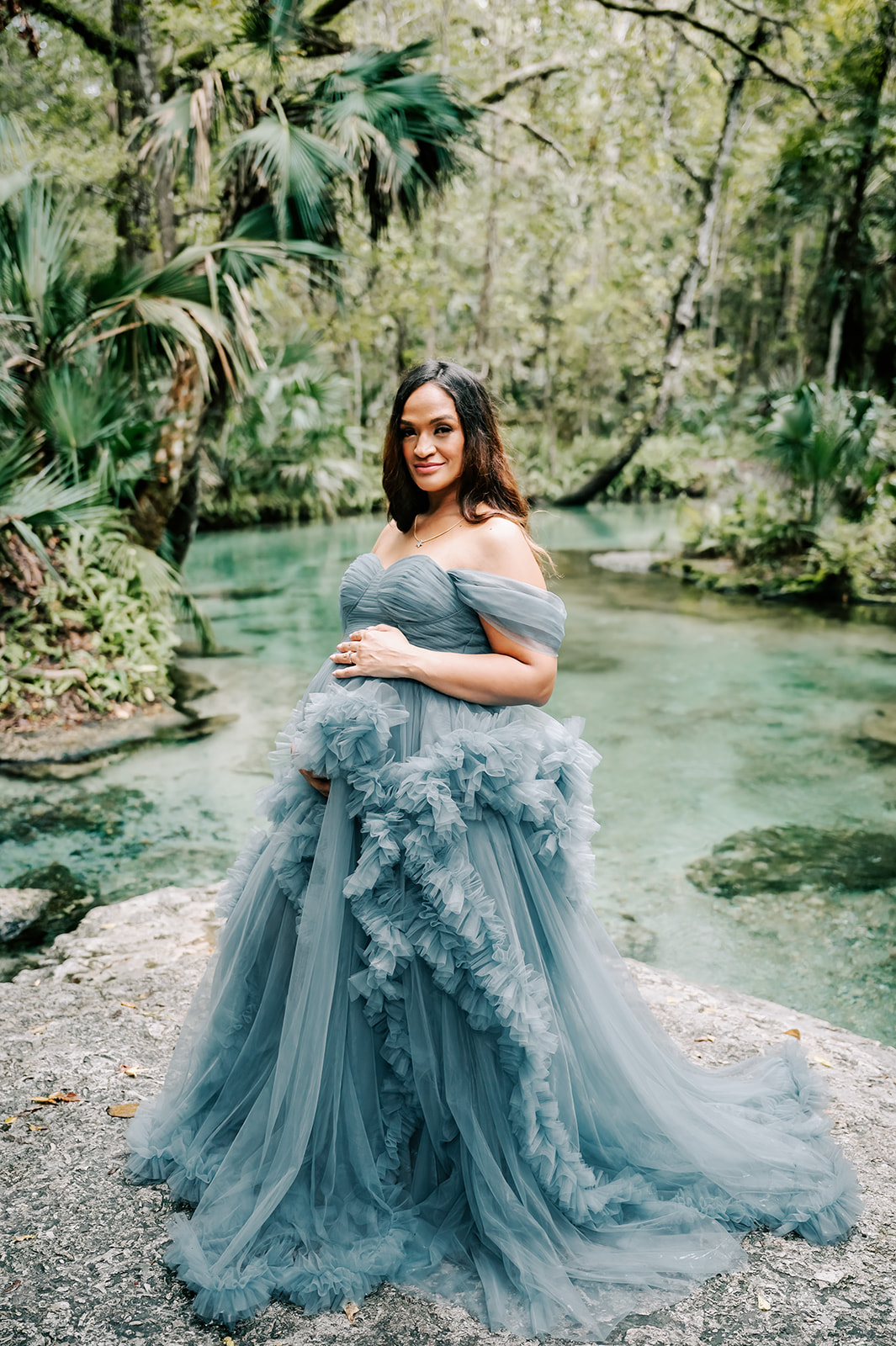 A mom to be in a long flowing tule maternity gown stands on a rock by a flowing clear river after visiting Moonshadow Medical Massage