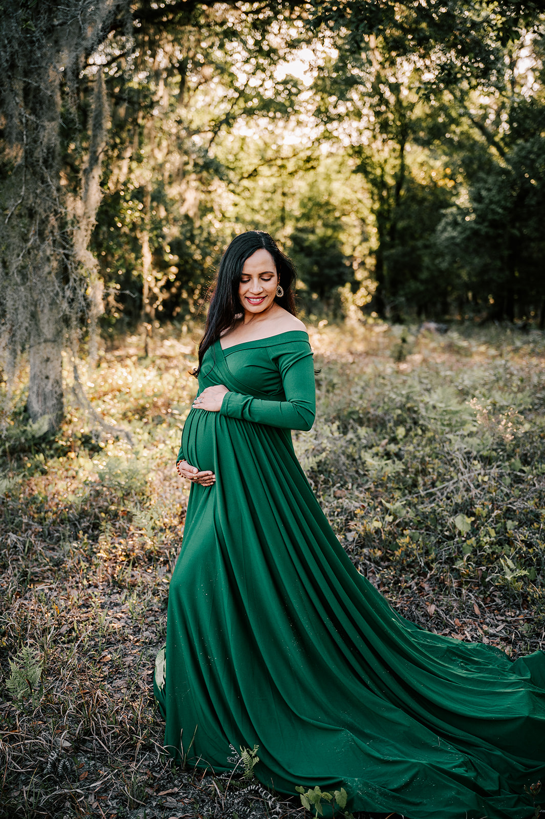 A mom to be smiles down her shoulder while standing in a forest at sunset in a green maternity gown with hands on her bump after visiting New Creation Birth Services