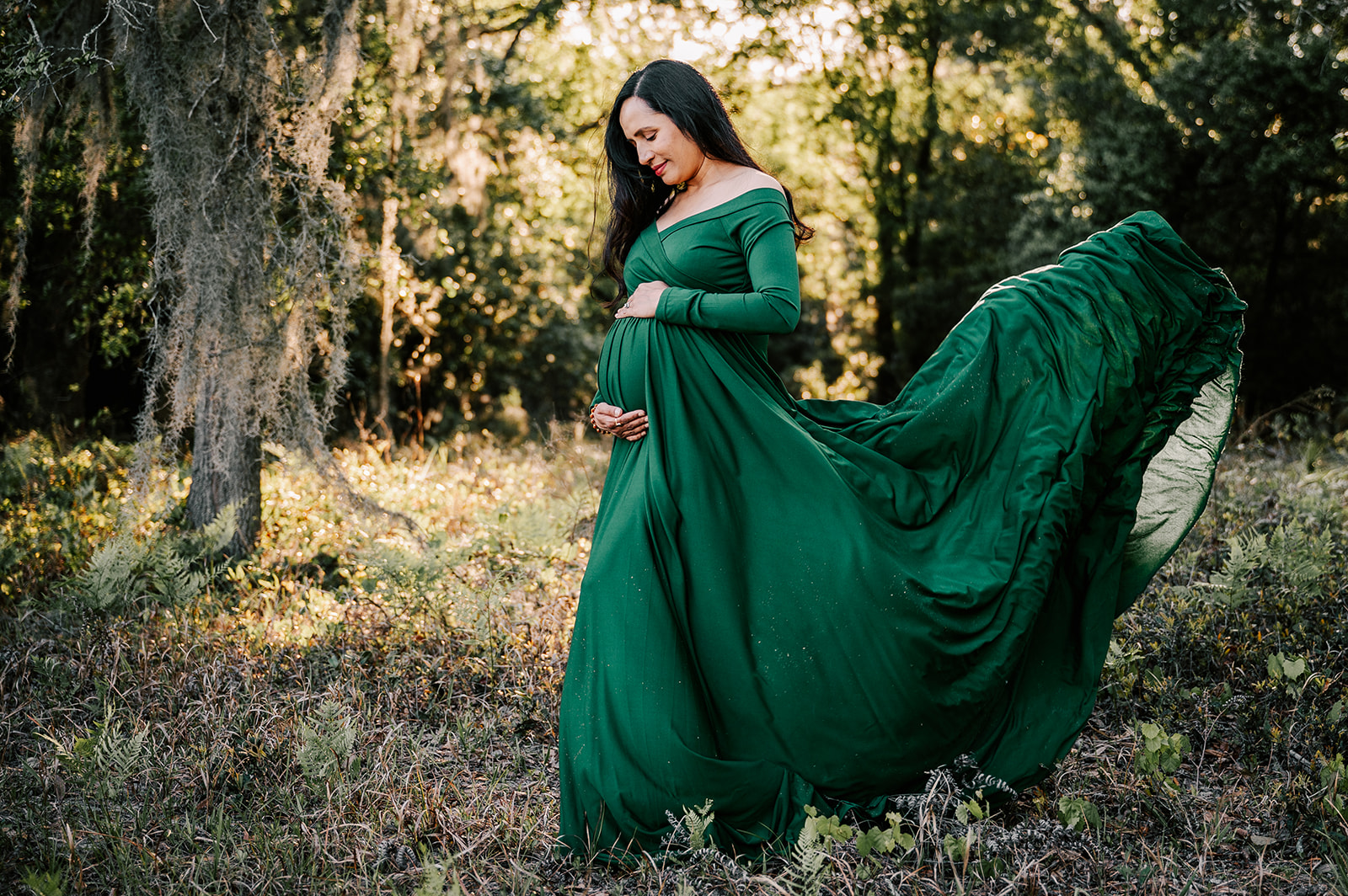 A mom to be in a long flowing green maternity gown smiles down at her bump in her hands at sunset in a forest after visiting New Creation Birth Services