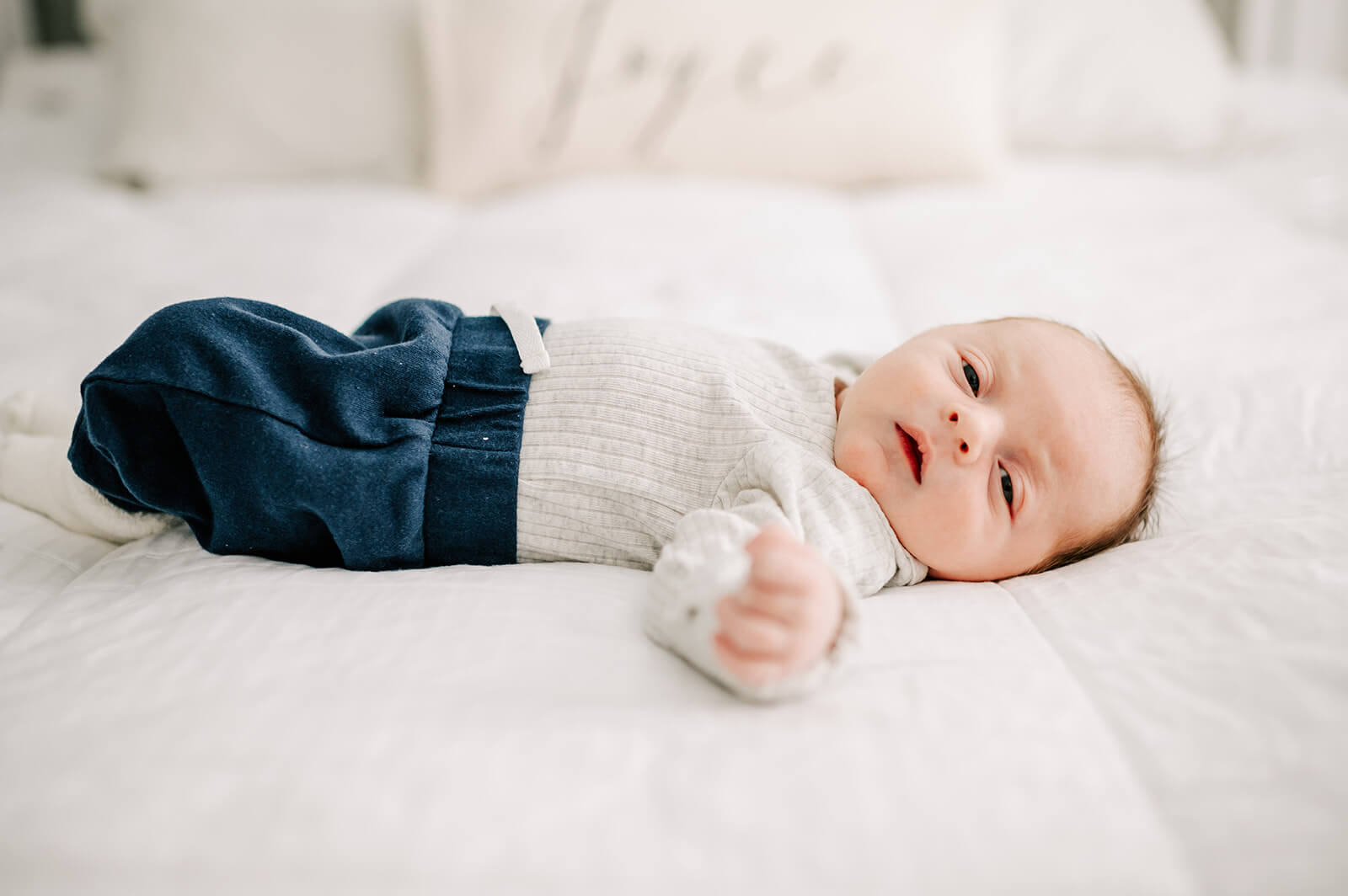 A newborn baby boy in a grey sweater and blue pants lays on a bed with eyes barely open