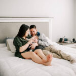 Happy new parents sit on their bed snuggling with their newborn baby in mom's arms after meeting novant health lactation consultants