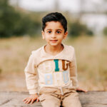 A toddler boy in a big bro shirt sits on a wooden bench in a park