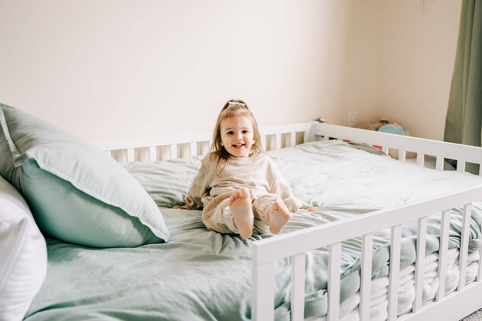 A toddler girl plays on a bed in her room with a big smile