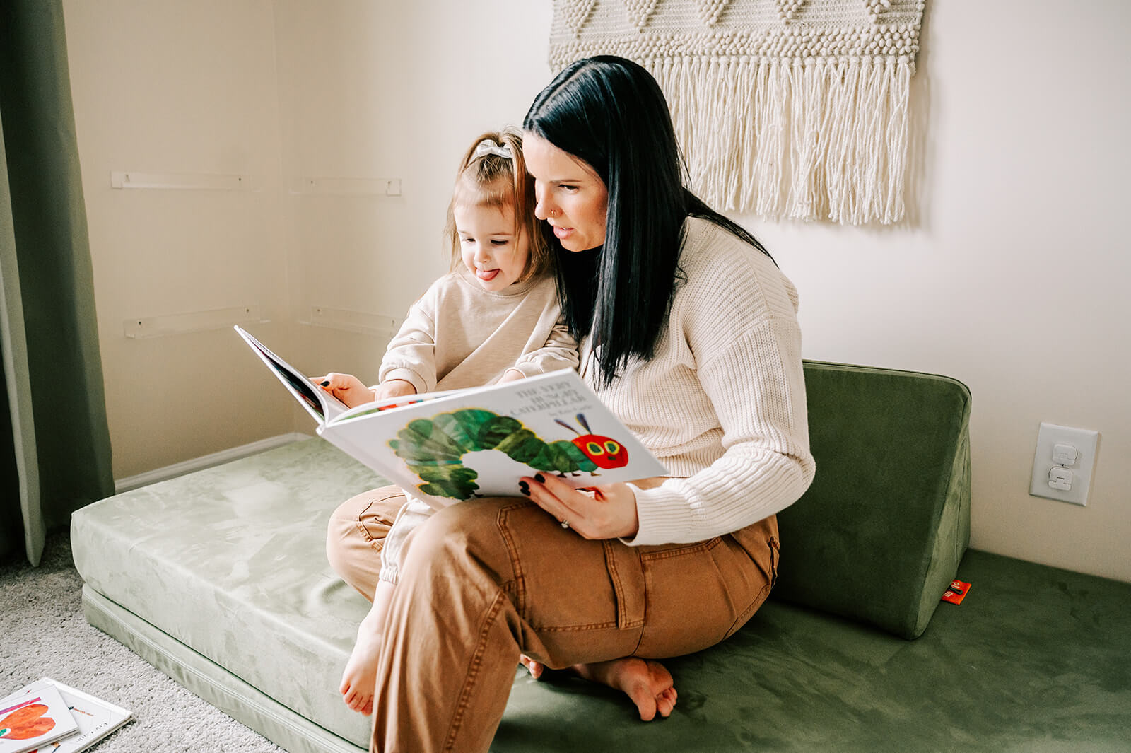 A mom in brown pants and a white sweater read a book with her toddler daughter in a play room by a window after visiting pottery barn kids durham