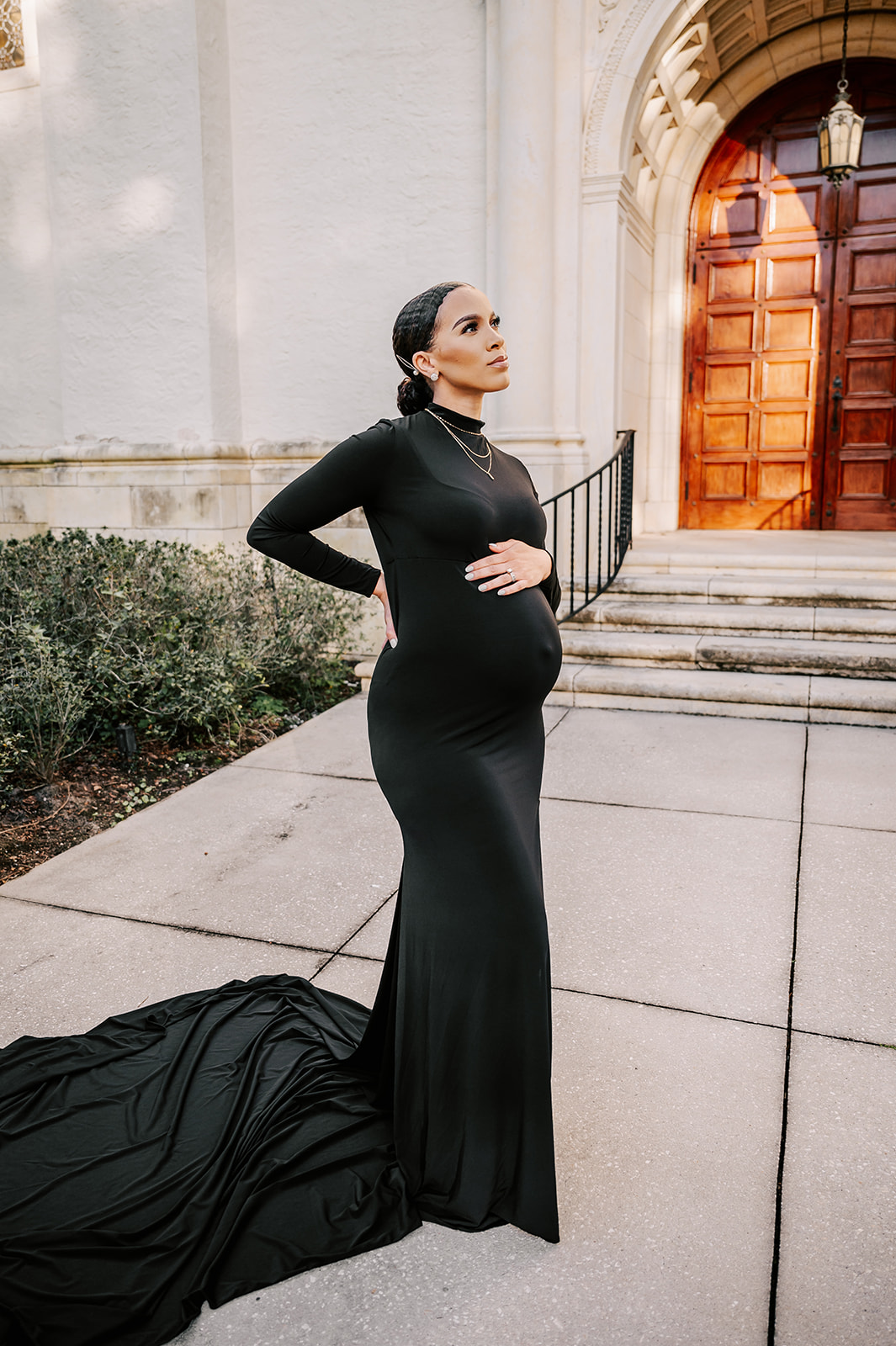 A mother to be in a long black maternity gown stands by a historical building with a hand on her bump