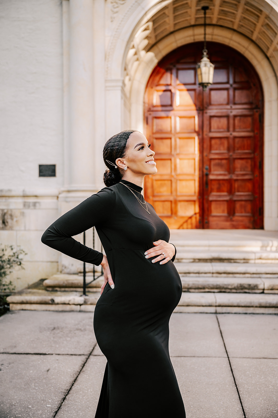 A mother to be smiles up to the sky while holding her bump and standing in front of large wooden doors of a church after visiting REX Birthing Center
