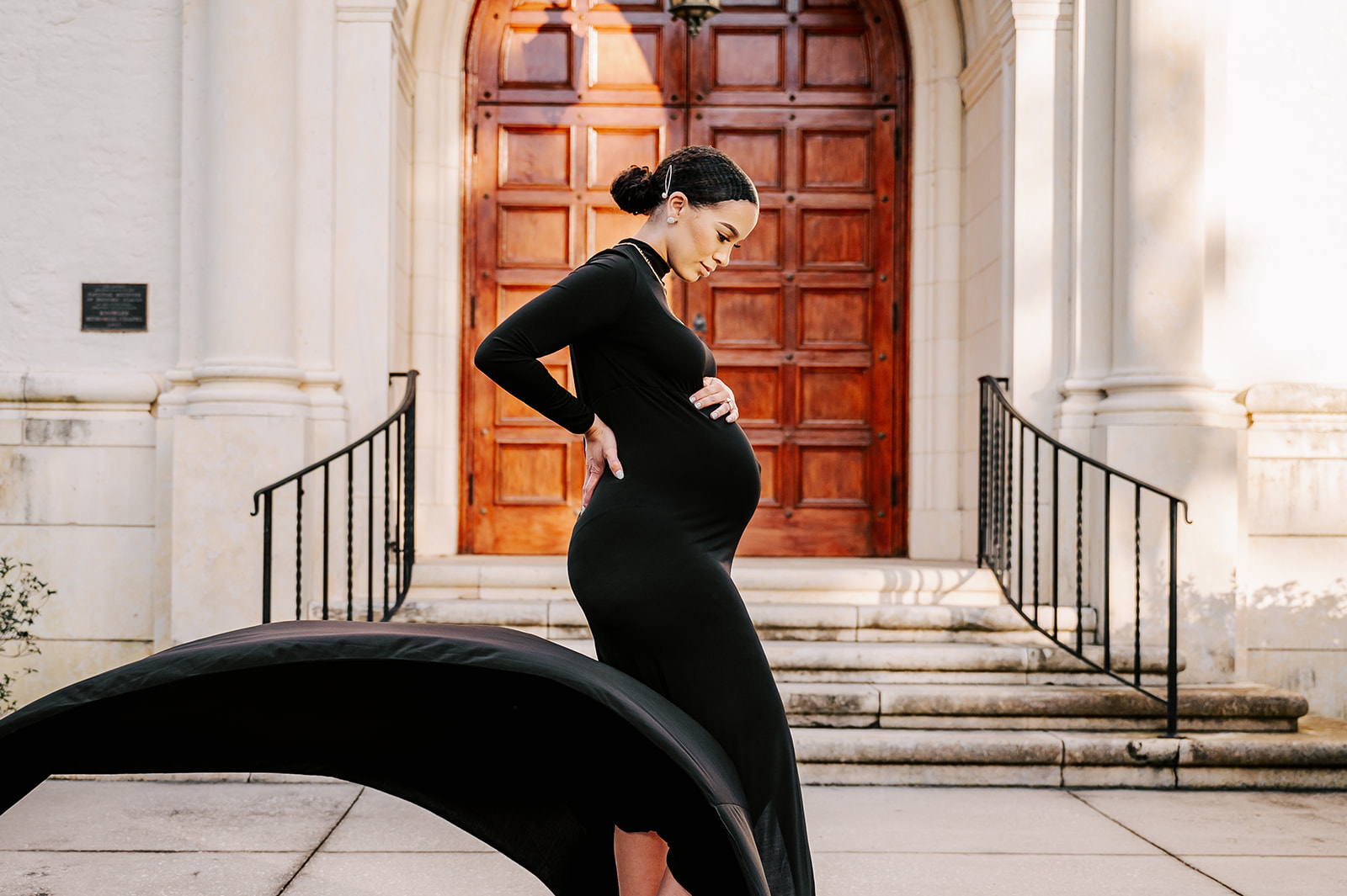 A mother to be in a long black maternity gown smiles downa t her bump in front of large ornate wooden doors after visiting REX Birthing Center