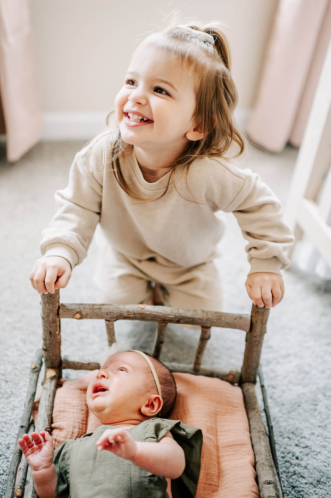A toddler girl in a tan sweater smiles overtop her newborn baby sister laying in a wooden stick bed in a nursery after visiting shower me with love charlotte