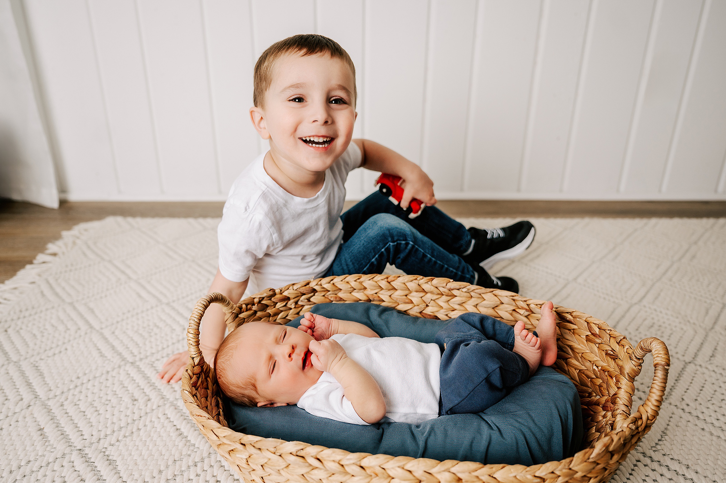 A happy toddler boy smiles while sitting with his sleeping newborn baby brother in a studio