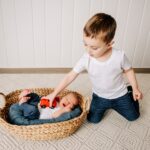 A toddler boy plays with a toy car on his sleeping newborn baby brother's belly on the floor of a studio after using Our Children's House