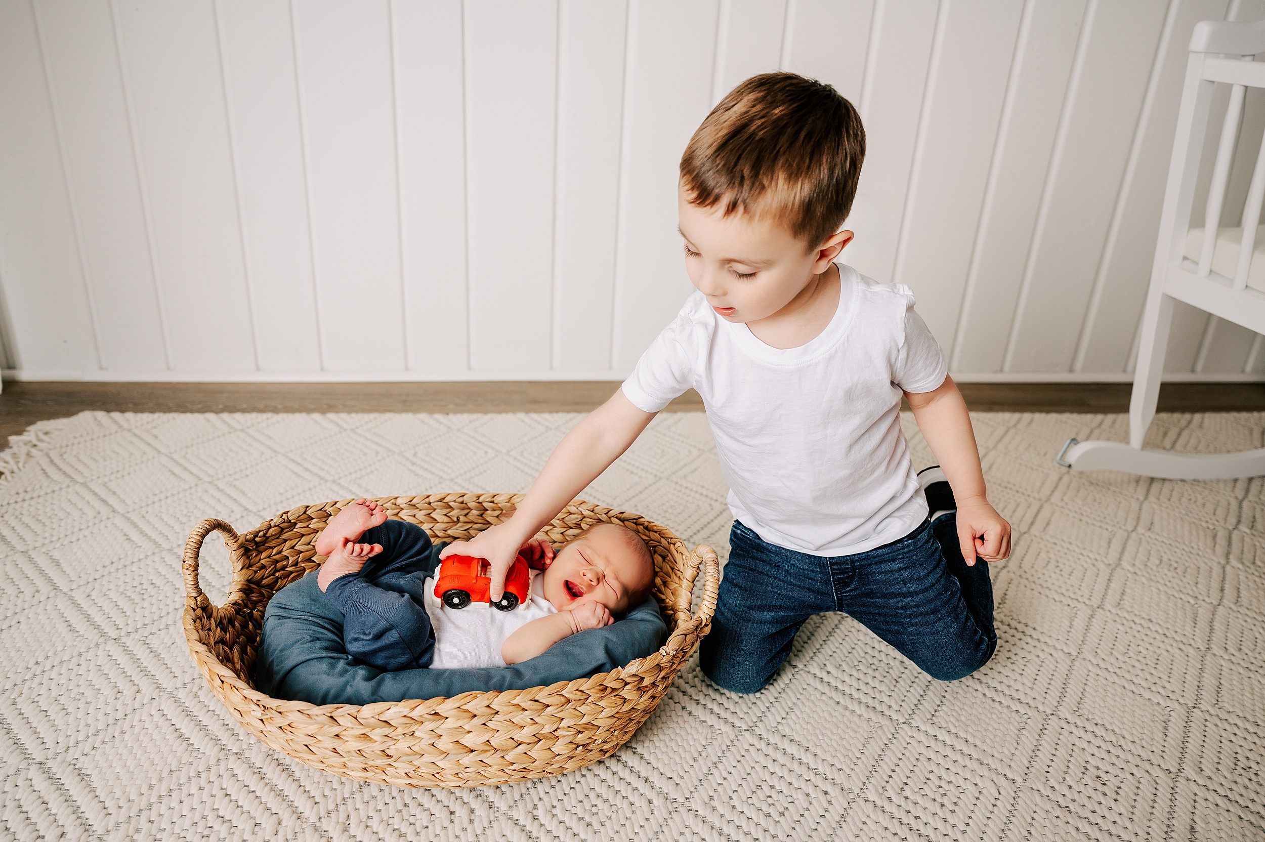 A toddler boy plays with a toy car on his sleeping newborn baby brother's belly on the floor of a studio after using Our Children's House