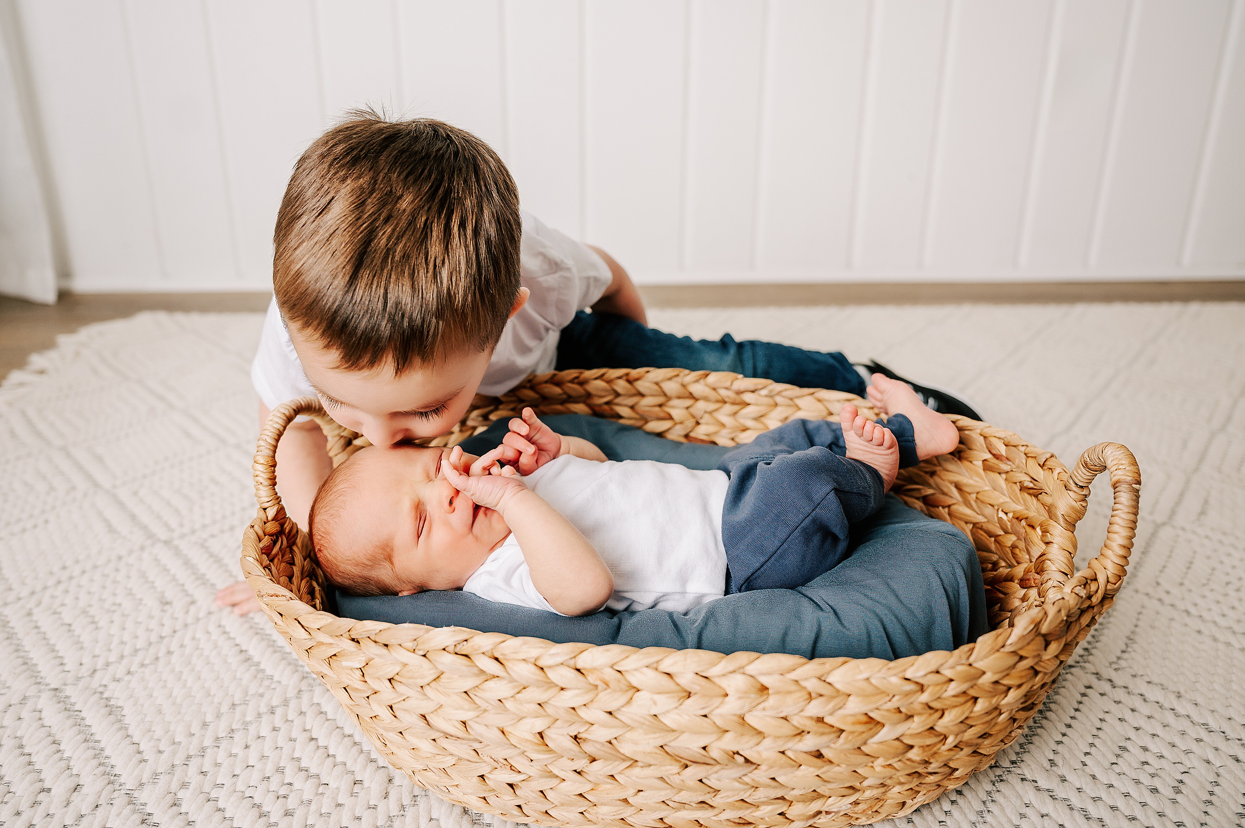 A young boy leans over and kisses his newborn baby brother laying in a woven basket after visiting Our Children's House