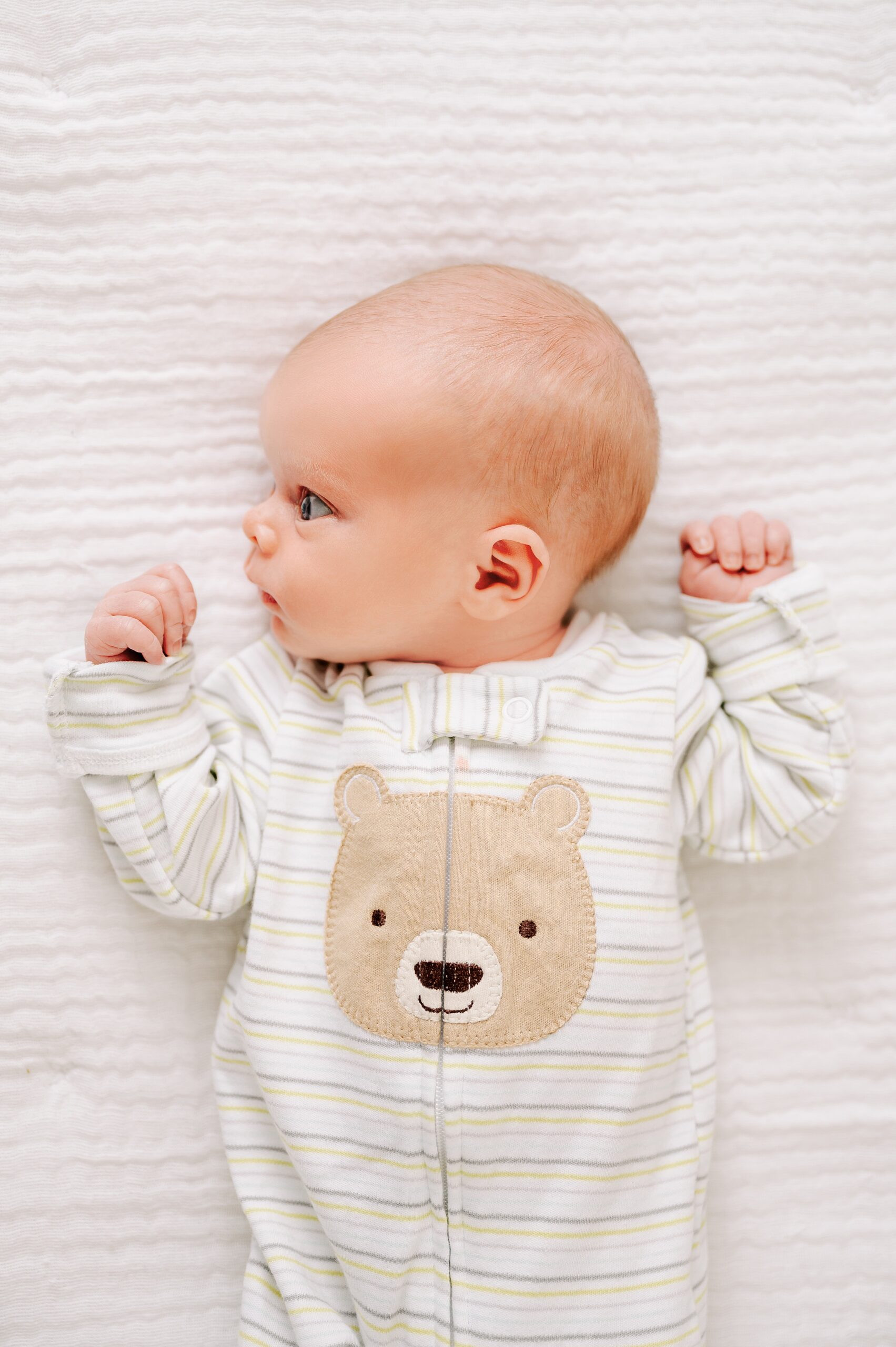 A newborn baby lays on a bed looking to the side in a bear onesie after leaving Wake Forest Birthing Center