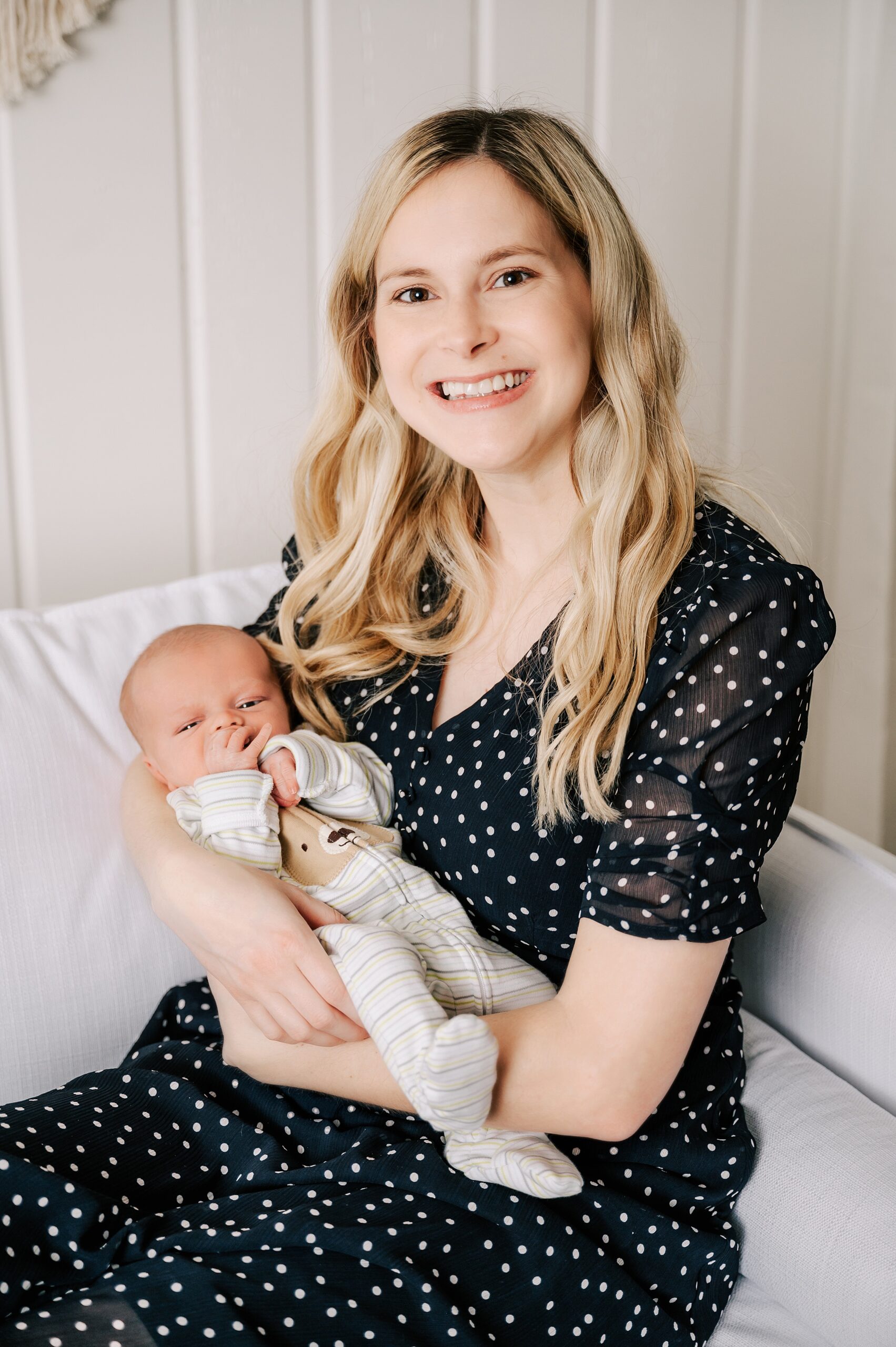 A new mom sits on a couch smiling while holding her newborn baby in a black polka dot dress thanks to Wake Forest Birthing Center