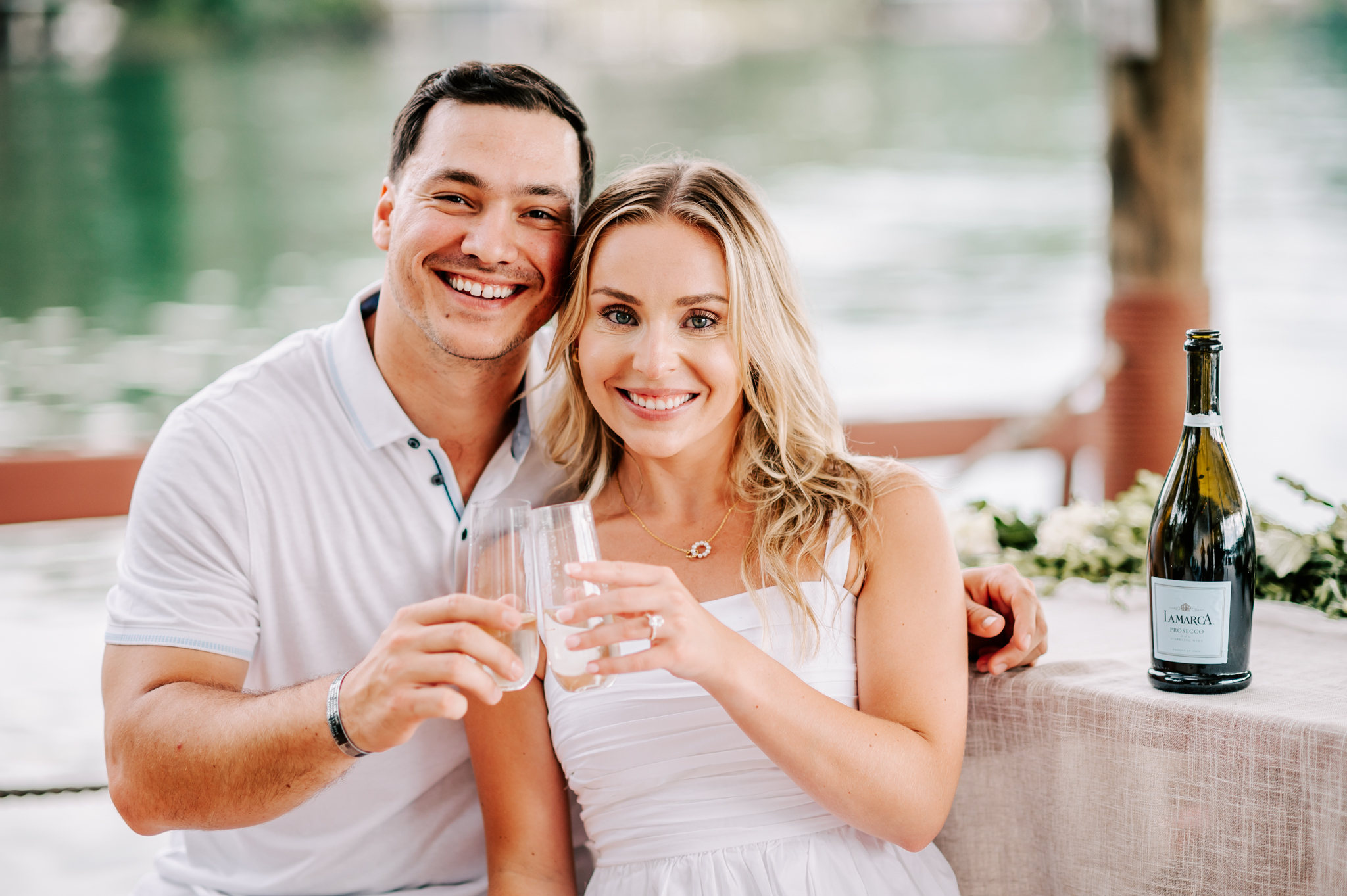 A happy engaged couple shares some champagne while sitting on a lake before visiting forsyth country club