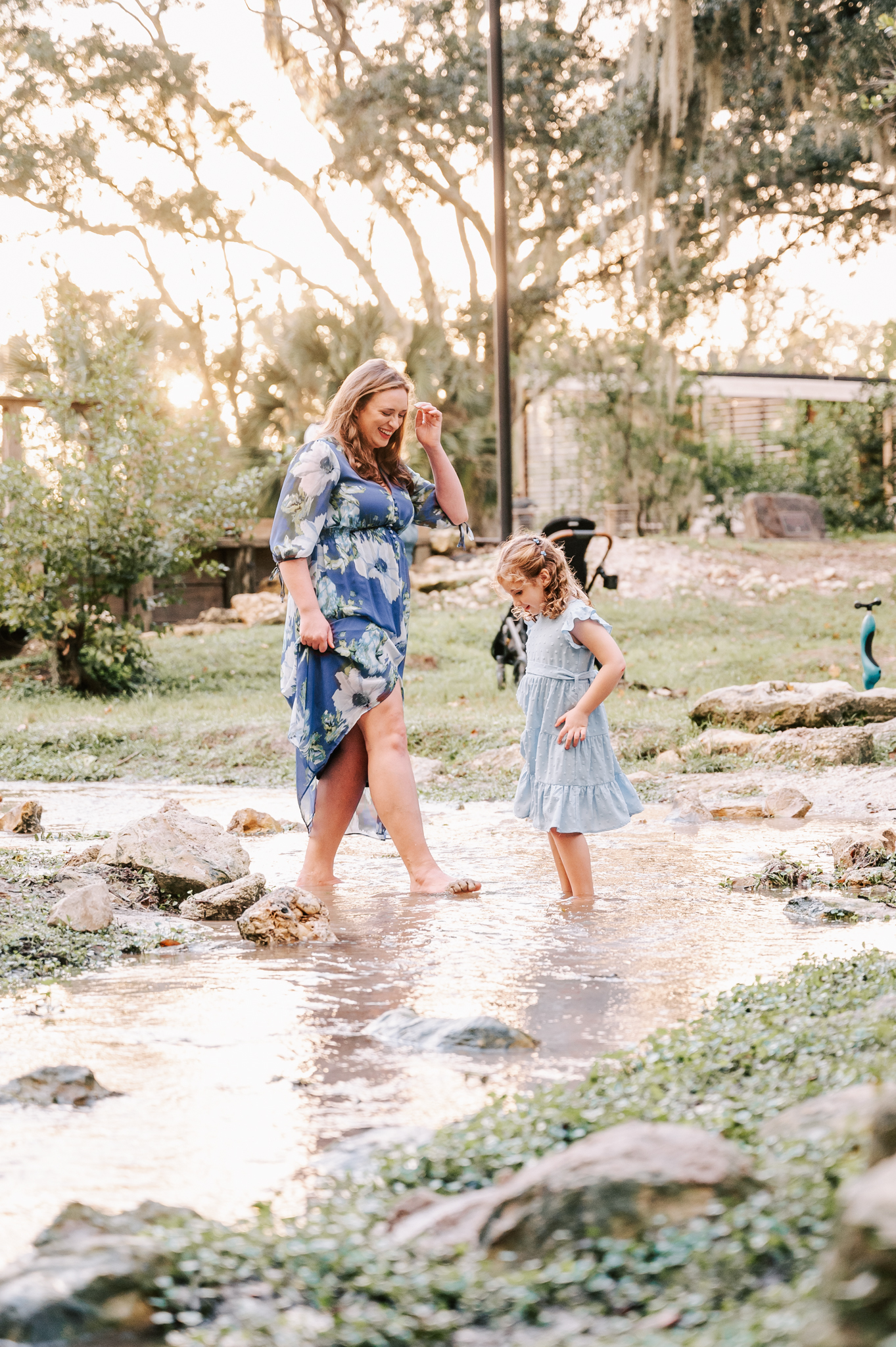 A mom and her young daughter in blue dresses play in a shallow creek at sunset