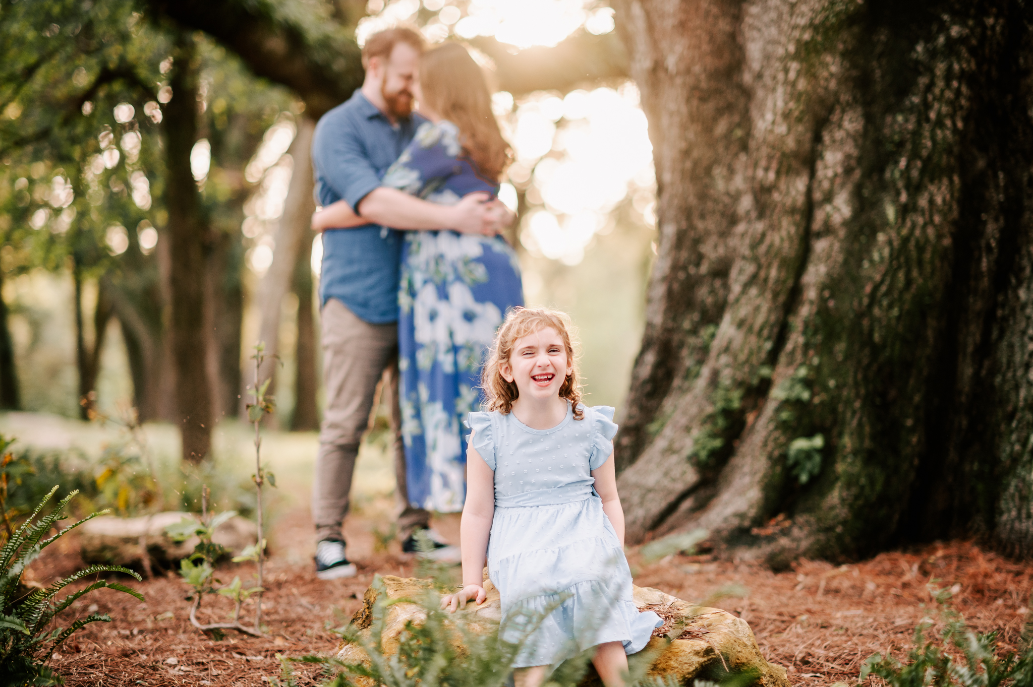 A young girl sits on a rock giggling while mom and dad snuggle at sunset