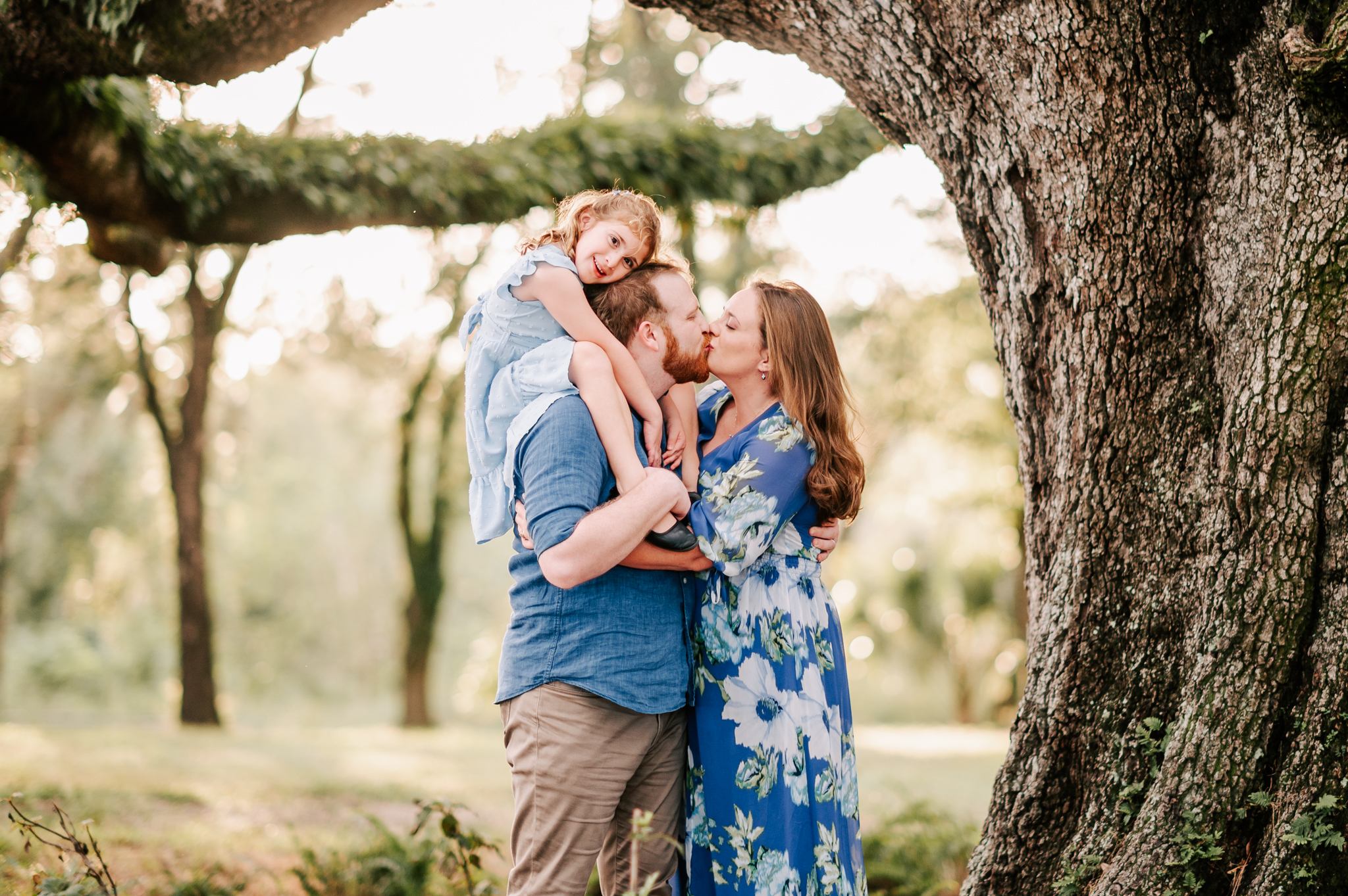 A young girl sits on dad's shoulders under a tree while he kisses mom