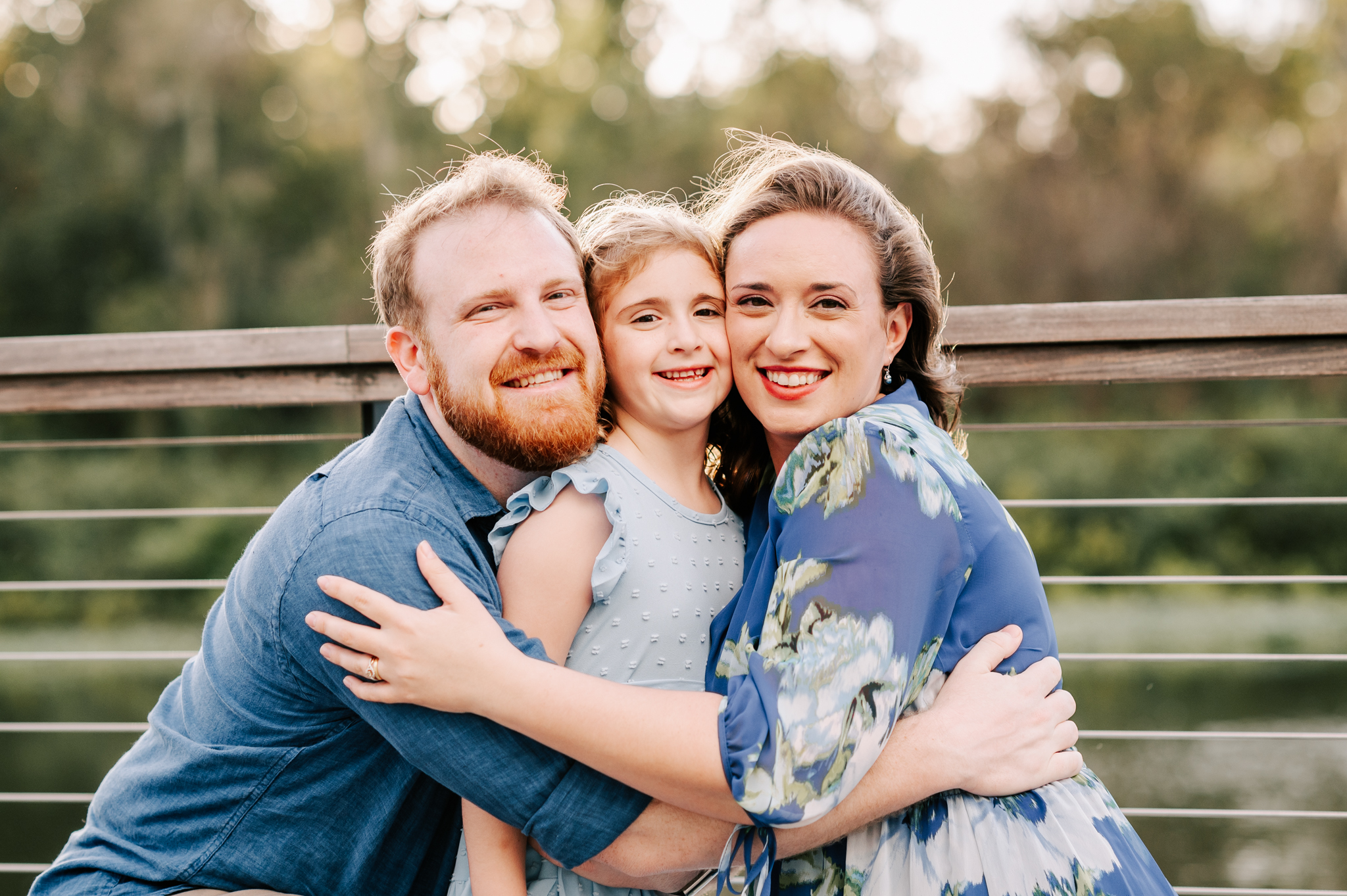 A young girl is hugged on both sides by mom and dad on a boardwalk at sunset