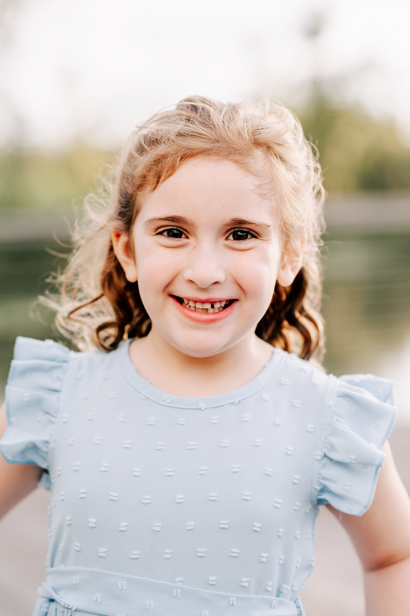 A young girl smiles big while exploring a park at sunset in a blue dress