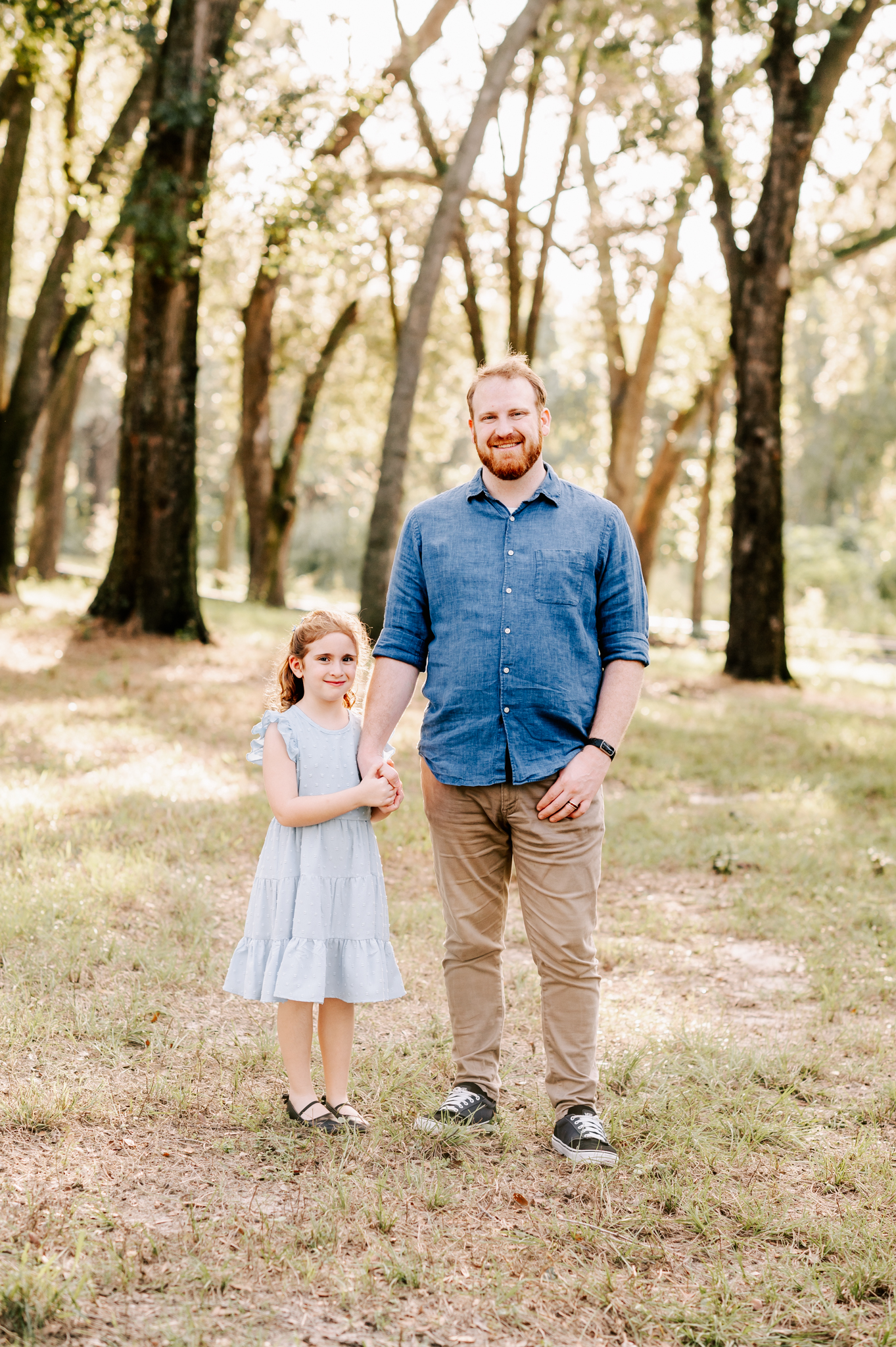 A happy dad stands in a park holding hands with his young daughter in a blue dress at sunset