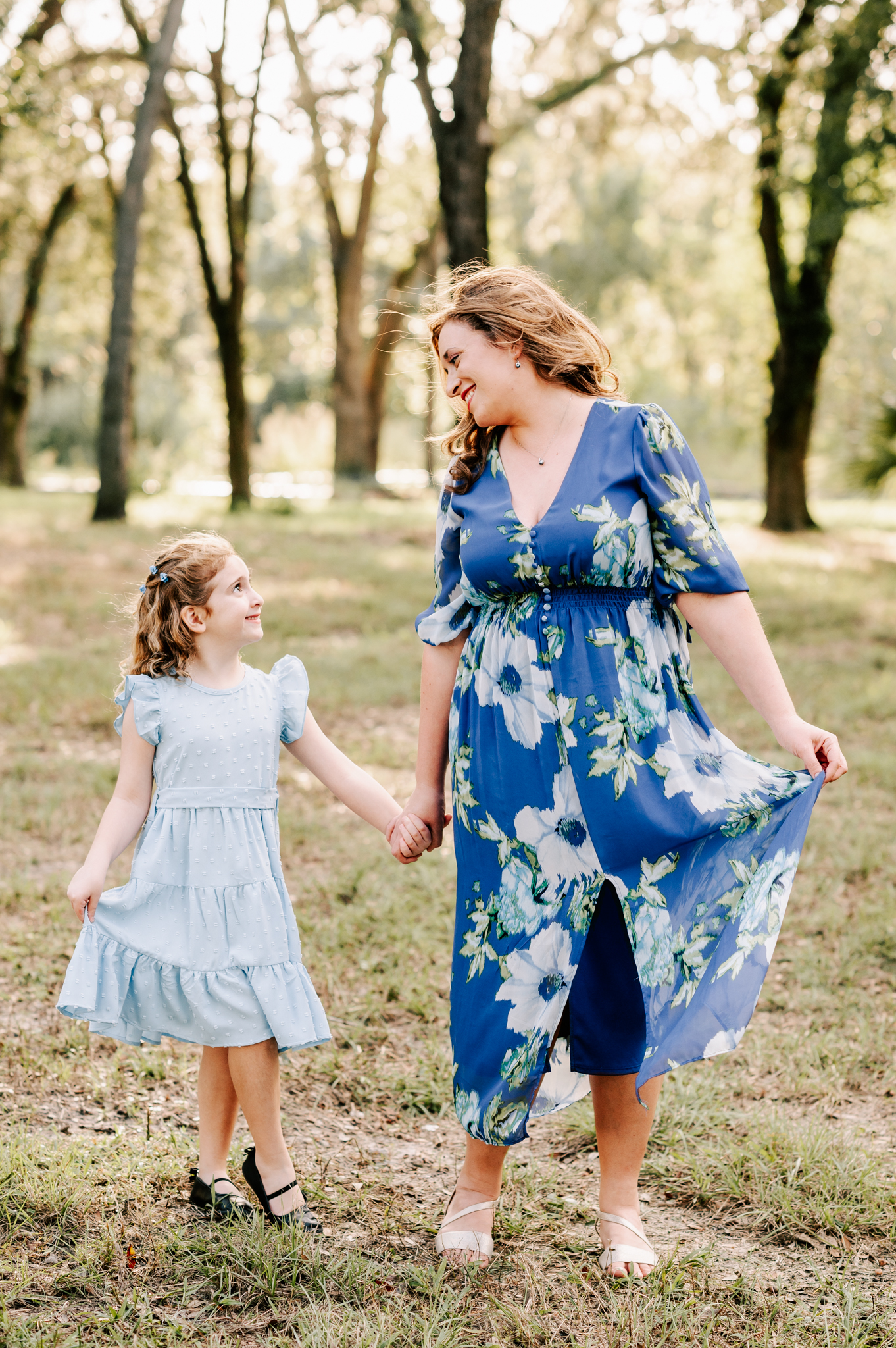 A mother and daughter in blue dresses walk through a park at sunset smiling at each other and holding hands before visiting north carolina fall festivals