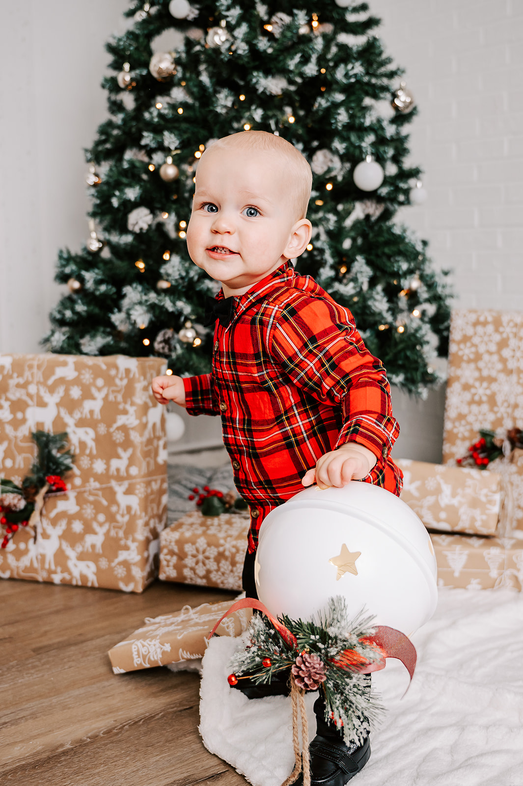 A young toddler boy plays with a christmas bell by the tree and presents in a plaid shirt and bowtie after visiting Piedmont Winterfest