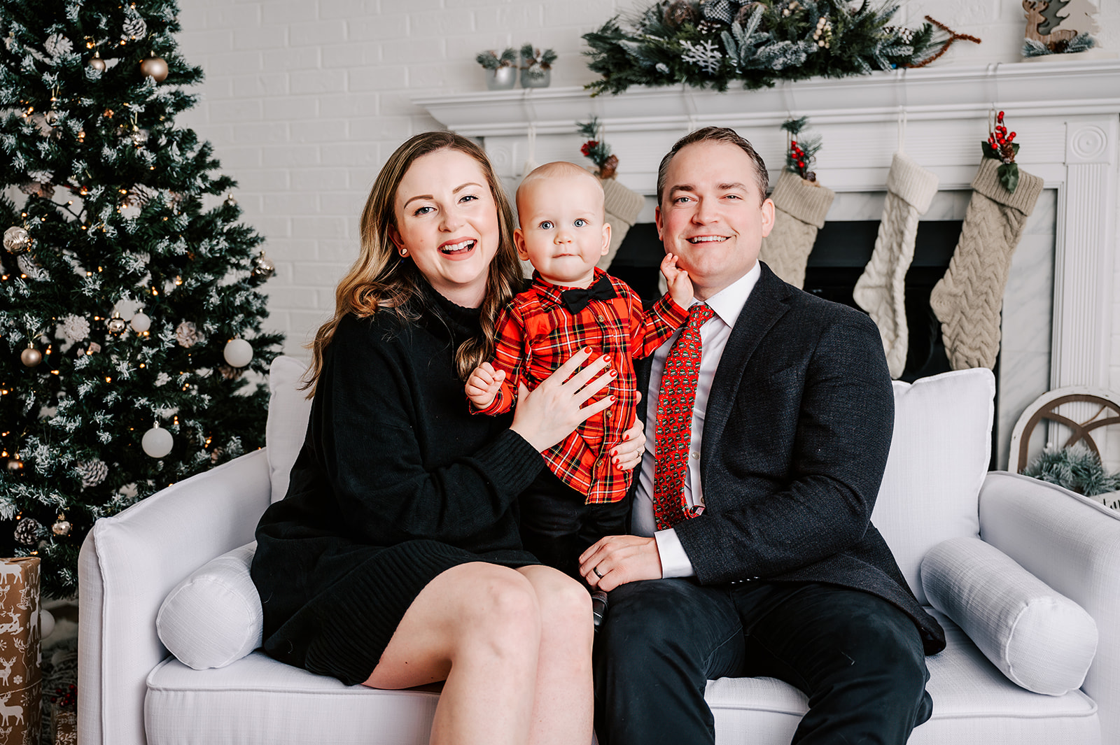 Happy mom and dad sit on a couch by the fireplace with their happy toddler son in their laps