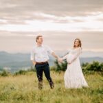 Happy expecting parents hike on a mountain pasture trail holding hands and smiling at sunset