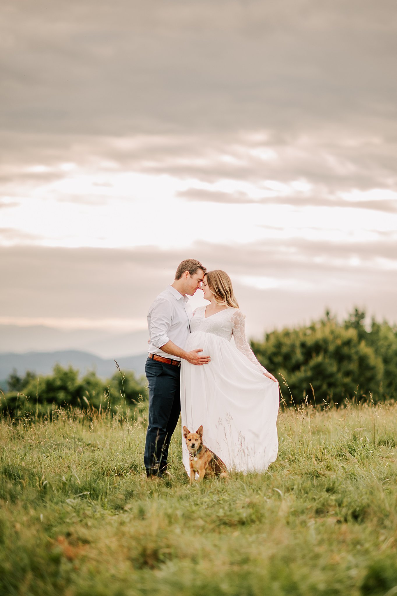 A pregnant woman in a white maternity gown stands in a mountain pasture while snuggling her husband with their dog sitting with them after some wakemed birthing classes