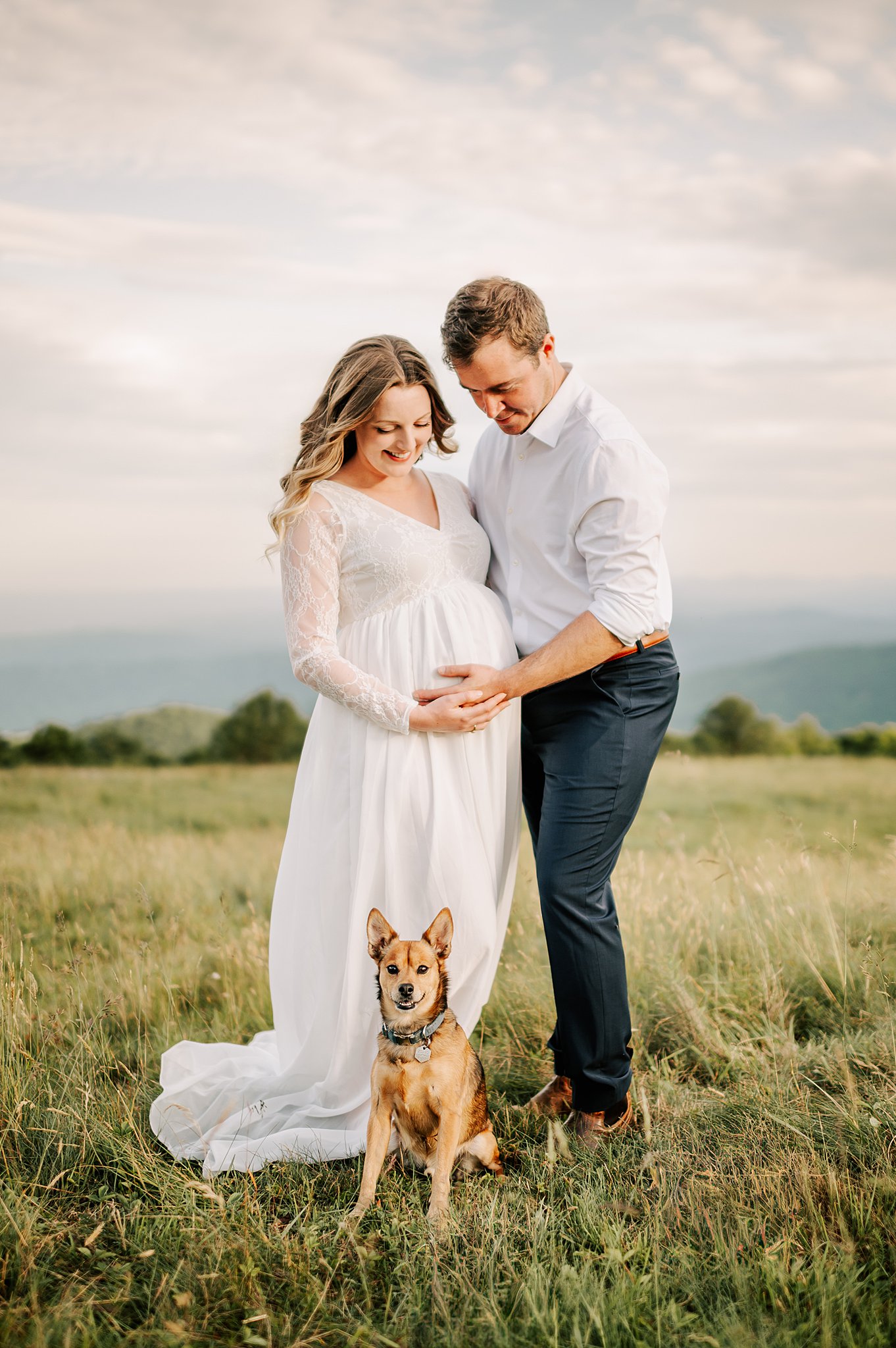 Happy expecting parents stand on a mountain pasture holding the bump with their small dog after some wakemed birthing classes