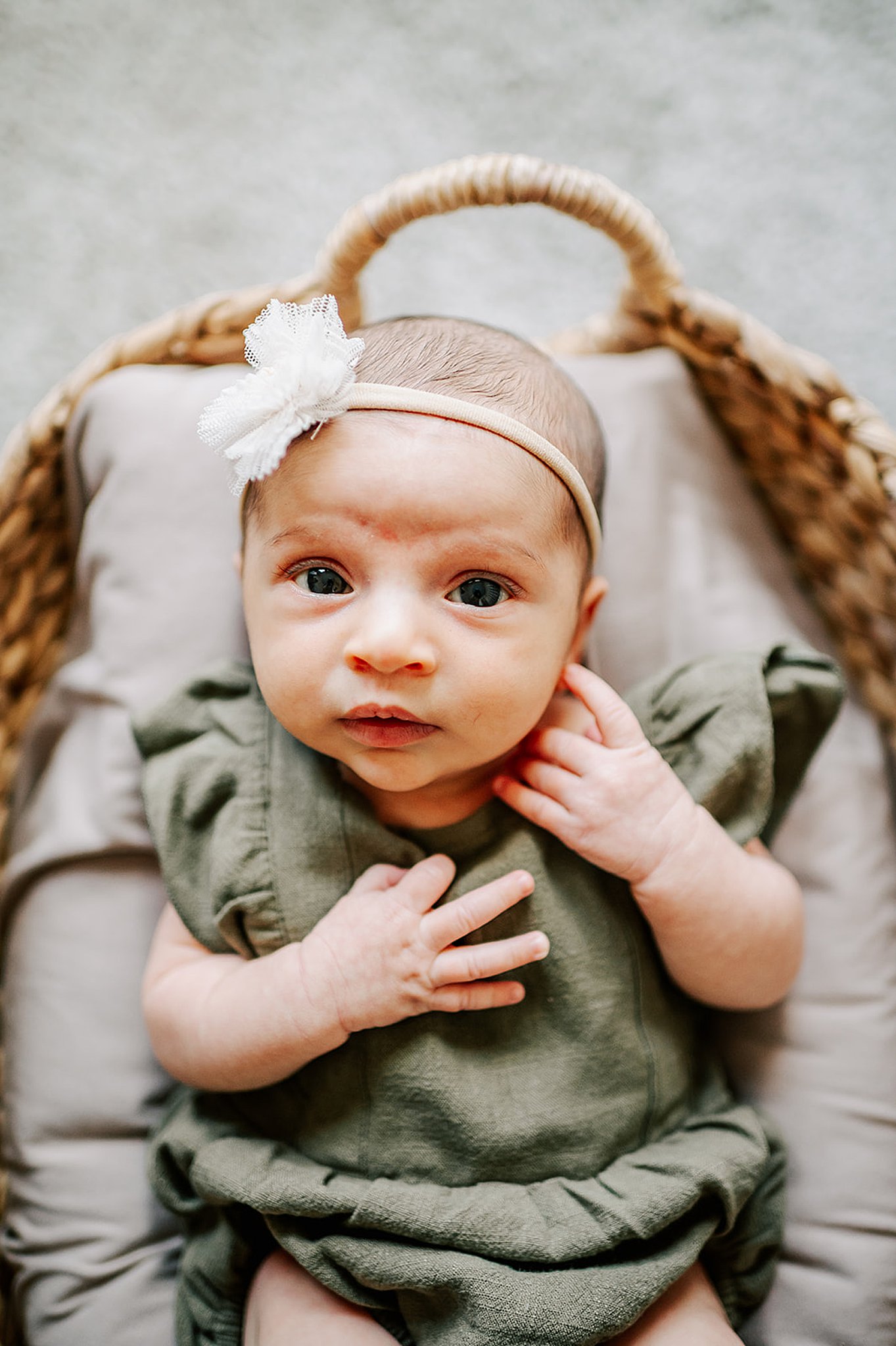 A newborn baby girl lays in a basket in a green dress and flower headband with eyes open