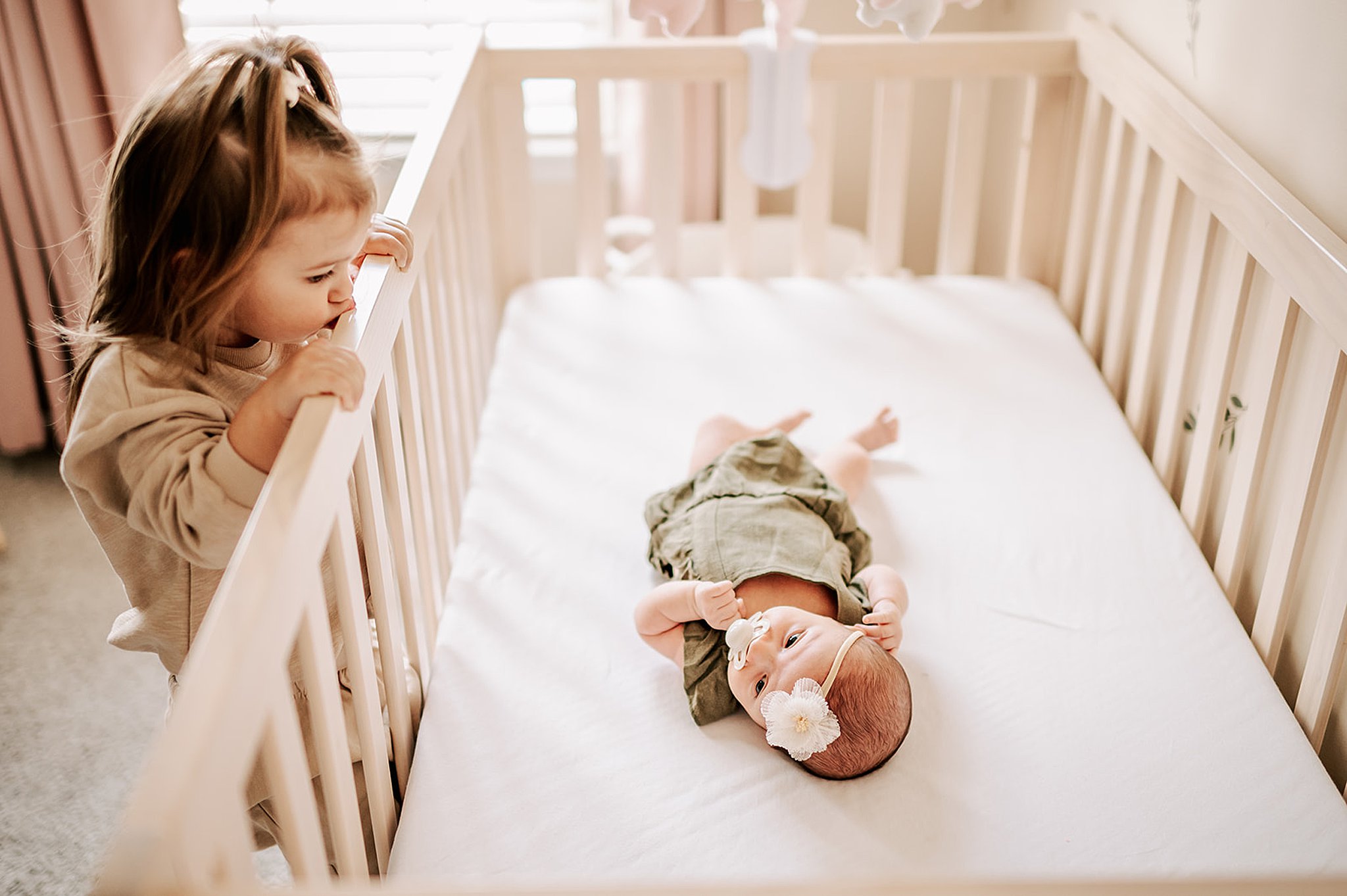 A toddler girl climbs up and looks down at her newborn baby sister laying in a crib